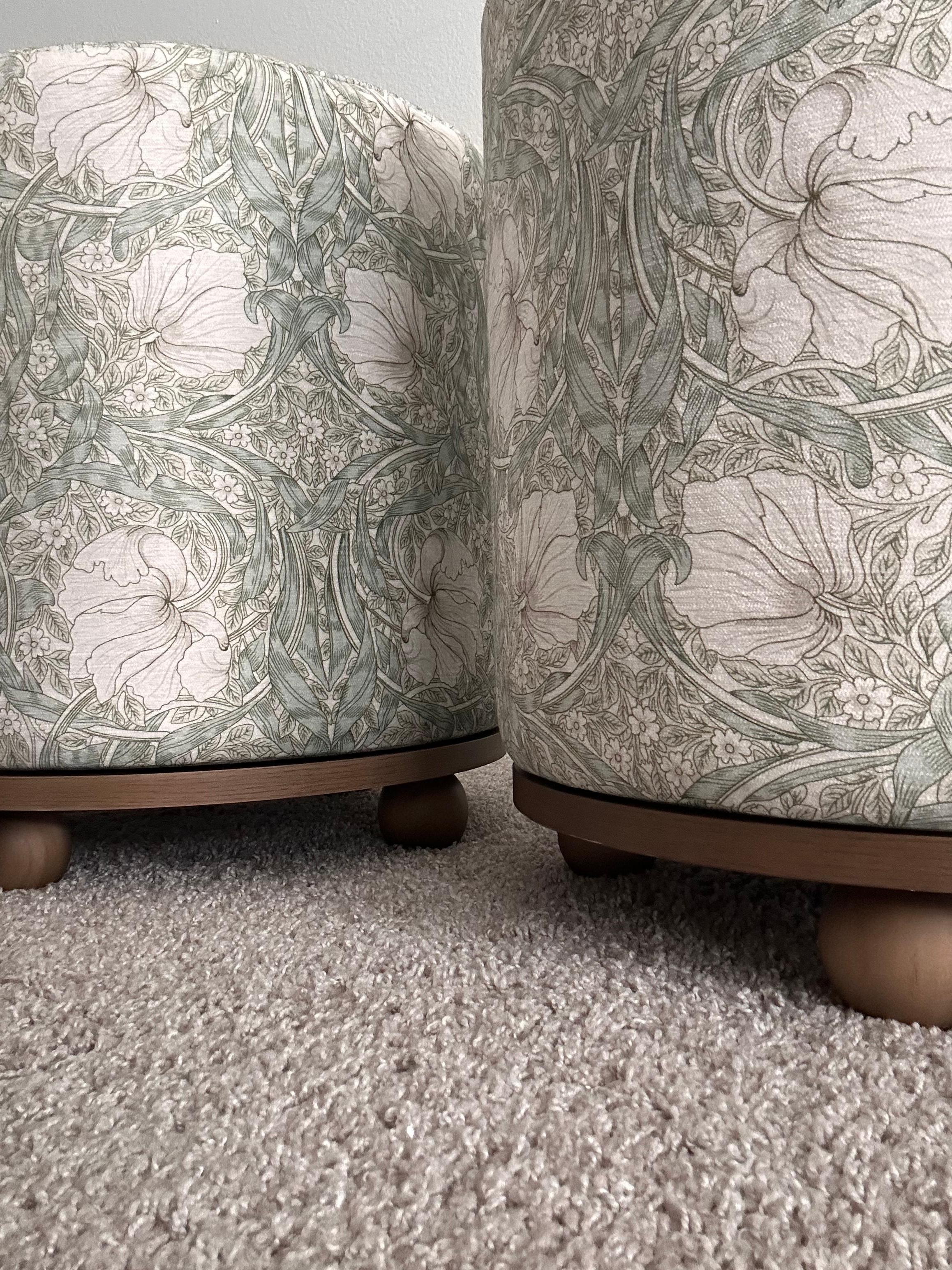 Pair of round sage green ottomans by Morris & Co. 
Stools are highlighted by the classic vintage floral-patterned fabric known to Morris & Co & supported by a swivel base with wooden ball feet, adding a unique and whimsical touch. 
They can be used