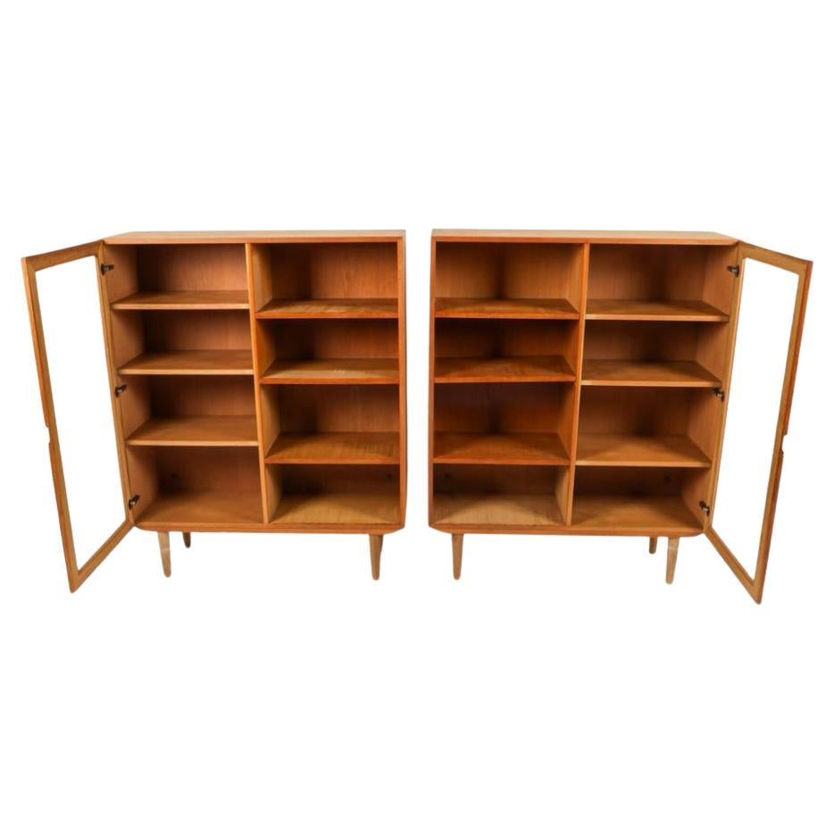 Pair of Scandinavian modern white oak bookcases with glass doors. Each book case or display case has (6) adjustable shelves with each bookcase having (1) glass door on opposite sides with concave handle. Each bookcase sits on (4) tapered oak legs.