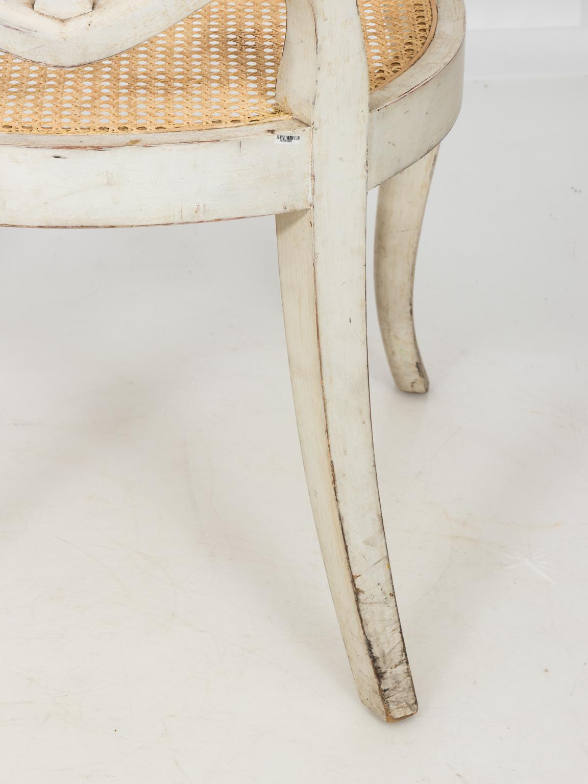White painted Scandinavian armchairs in the neoclassical style with ribbon back, cane seating, and cabriole legs. Please note of wear to the arms and legs due to daily use causing minor paint loss. The seat rail of one chair also has minor paint