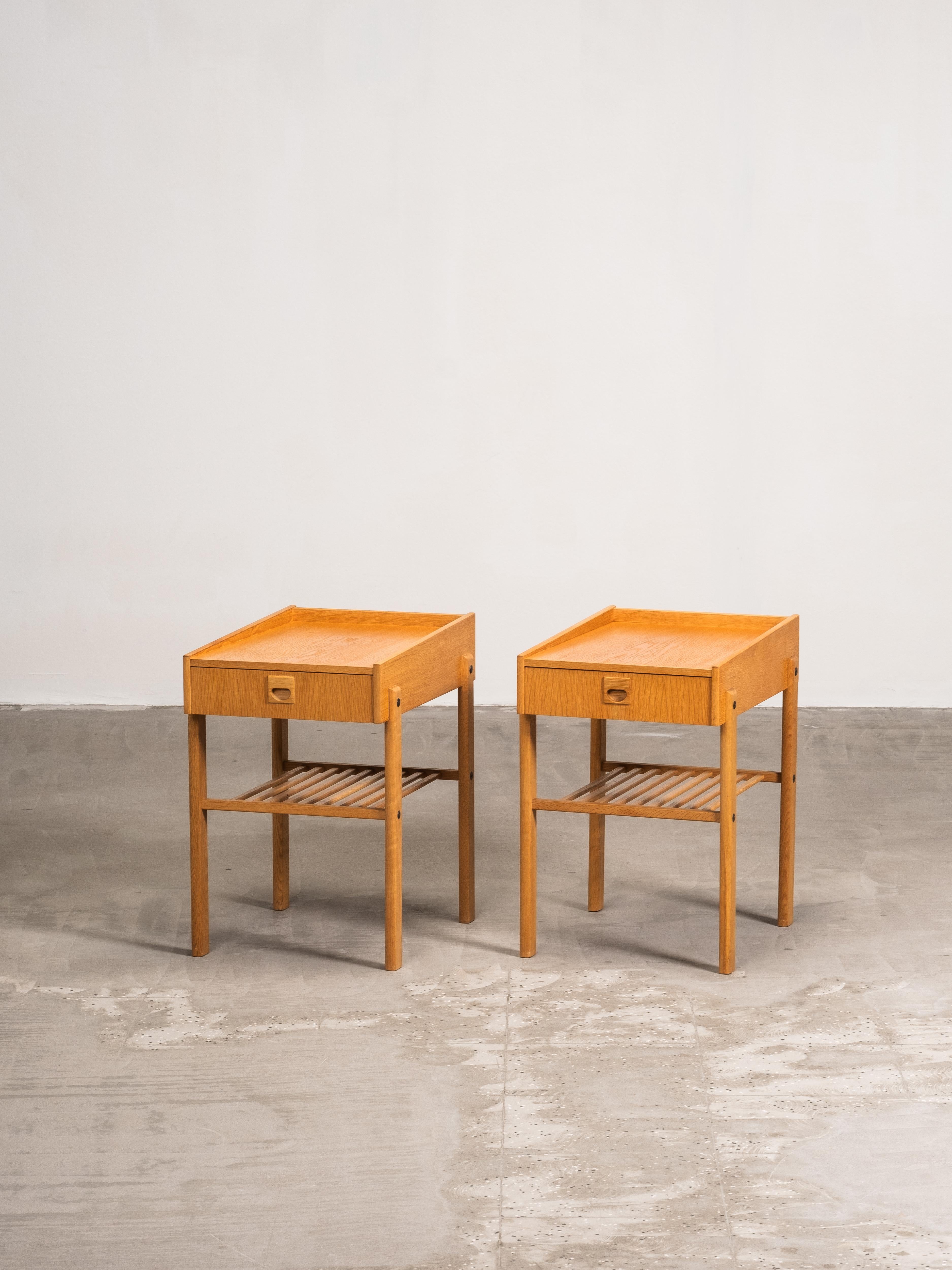 A pair of Swedish mid-century bedside tables in solid oak. Each table features a drawer with recessed handle, raised edges along the top surface, and a slatted lower shelf for books or magazines. The proportions are light and balanced, with square