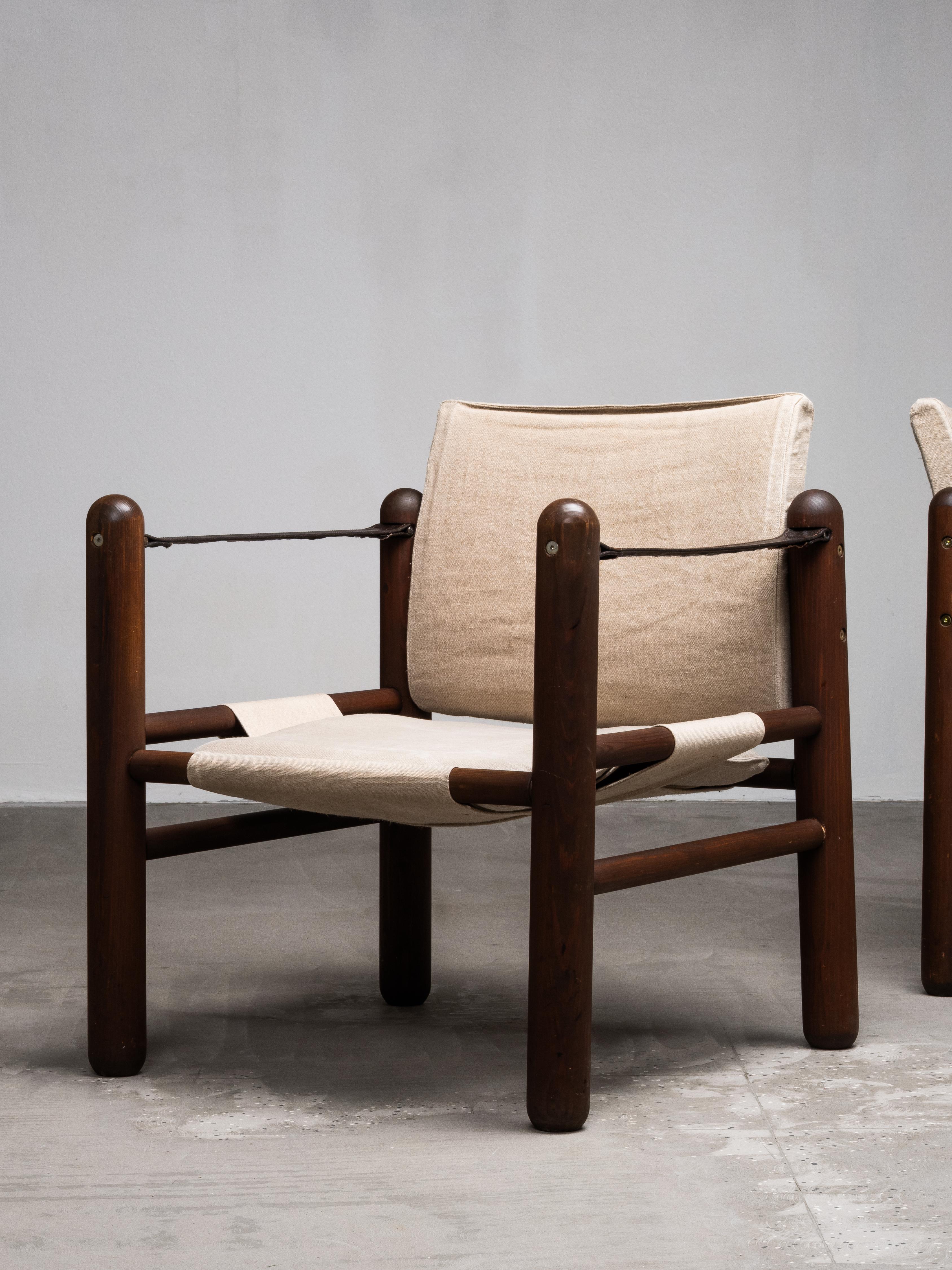 Paire de chaises safari des années 1970, en pin teinté, avec des élingues en lin naturel et des coussins de dossier amovibles. Le design utilise des poteaux en pin épais et arrondis, reliés par des châssis horizontaux, créant ainsi un cadre robuste