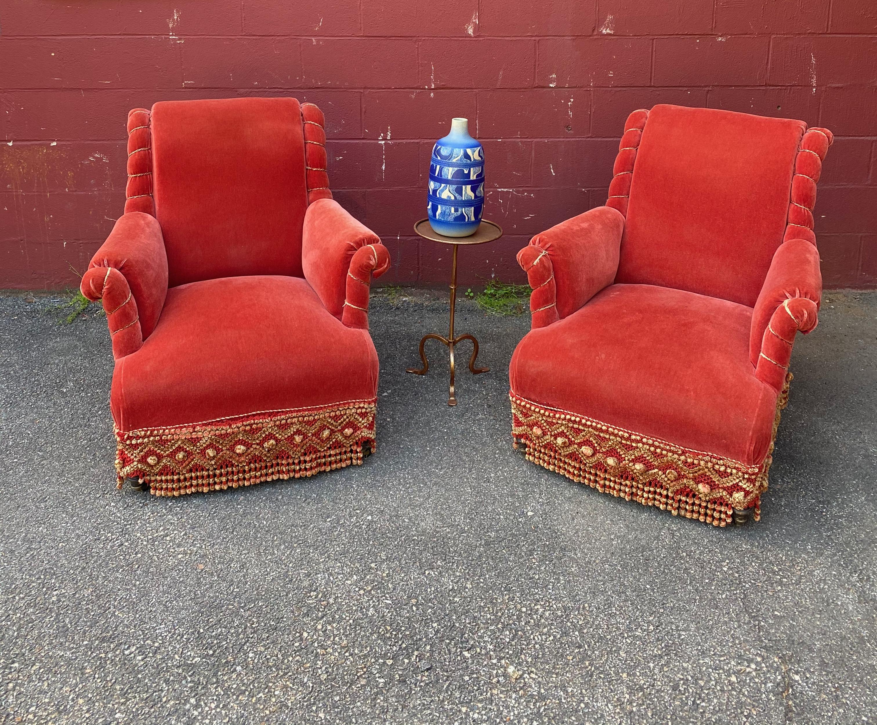 An elaborately upholstered pair of French armchairs with scrolled backs. The vintage fabric is a rich orange / persimmon velvet that is accented by a braided rope along the arms and sides and is trimmed in a braided and tasseled fringe. The fabric