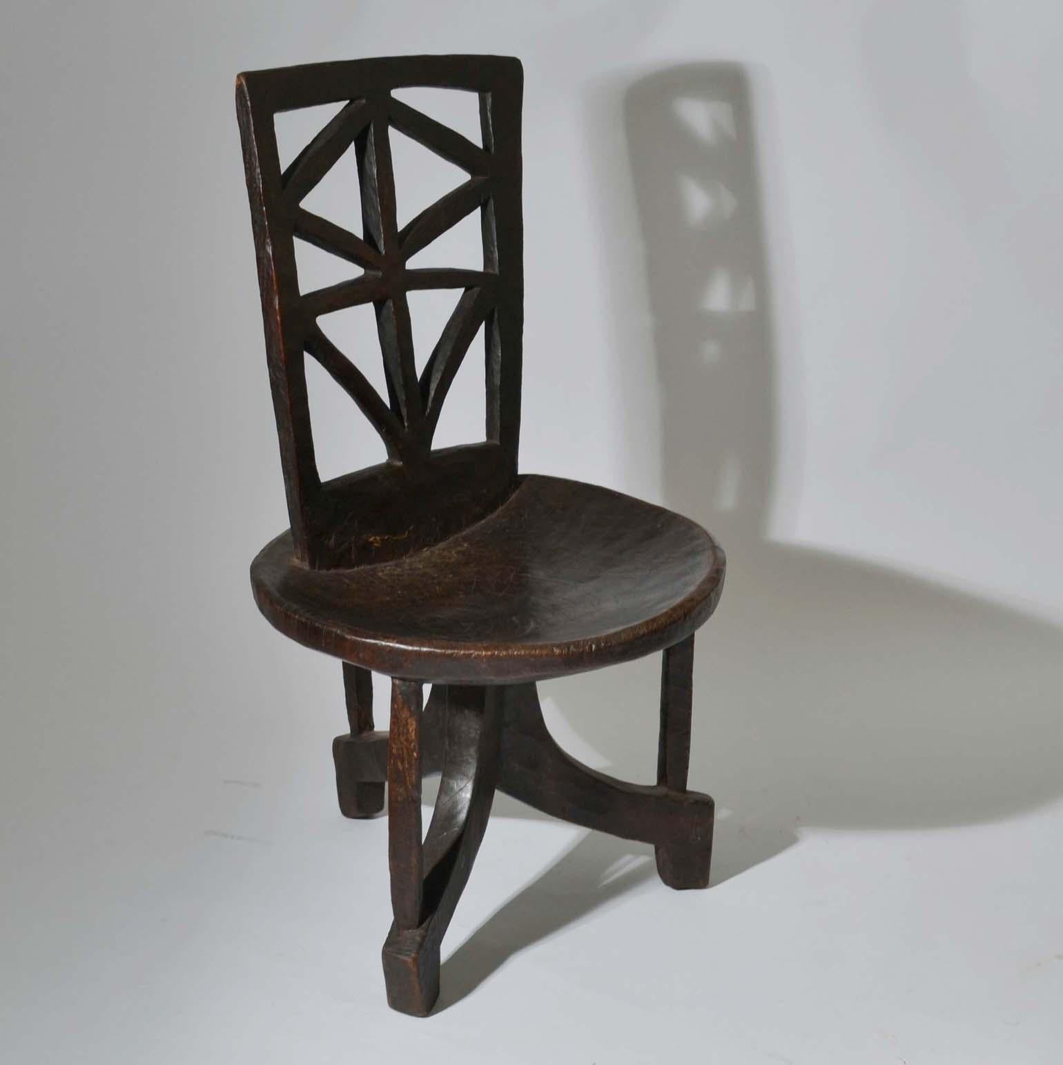 Two 1960s geometric high back Ethiopian chairs and a stool are all hand carved from one piece of wood. Circular, slightly concave seat on curved tripod base rectangular, backrest with geometric openwork carving and the dark wood has a fabulous shiny