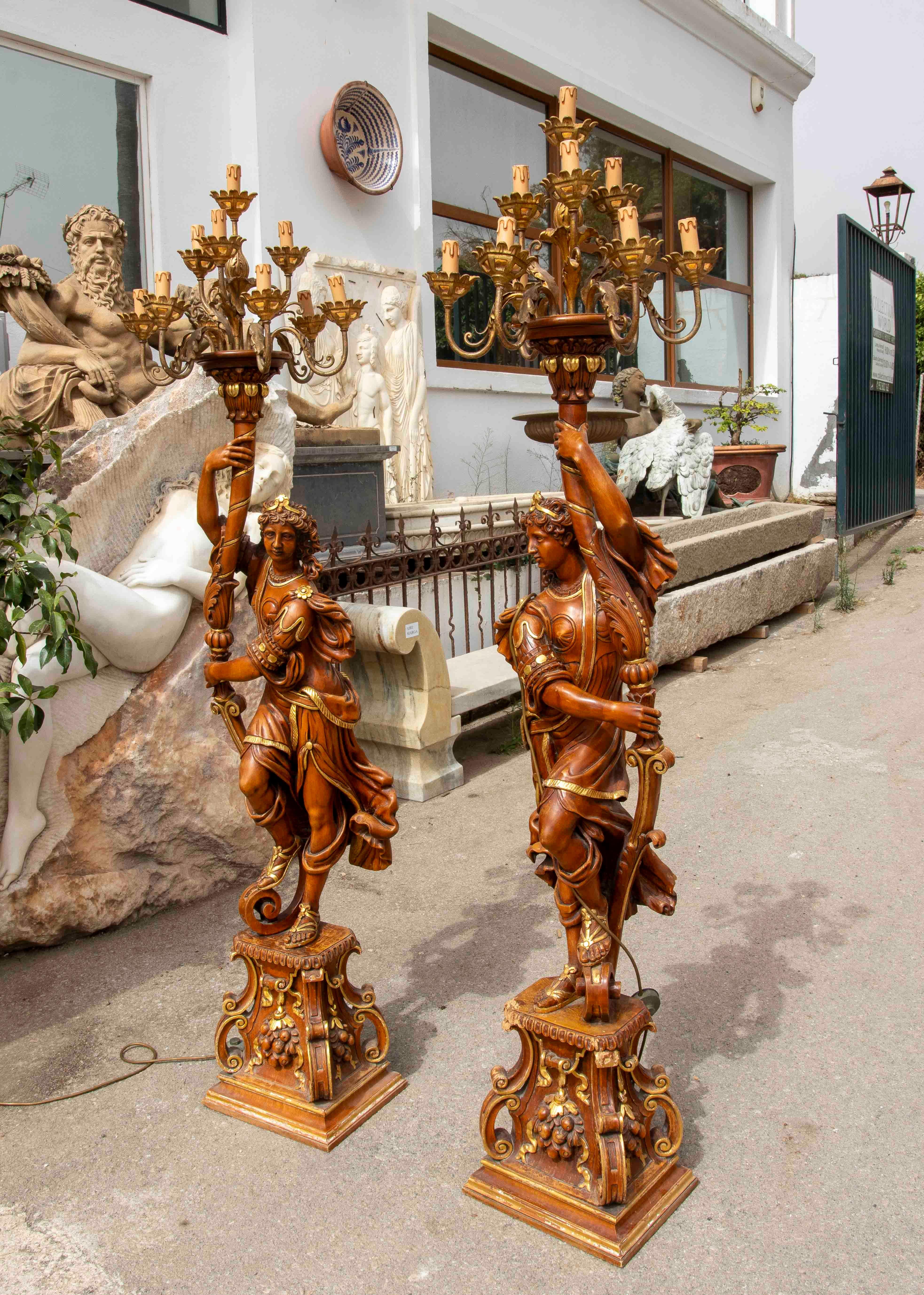 A stunning pair of sculptural candelabras made of carved and polychrome wood, depicting dynamic allegorical figures in classical poses. Each figure holds an elaborate arm culminating in a multi-candelabra with gilt ends, currently electrified for