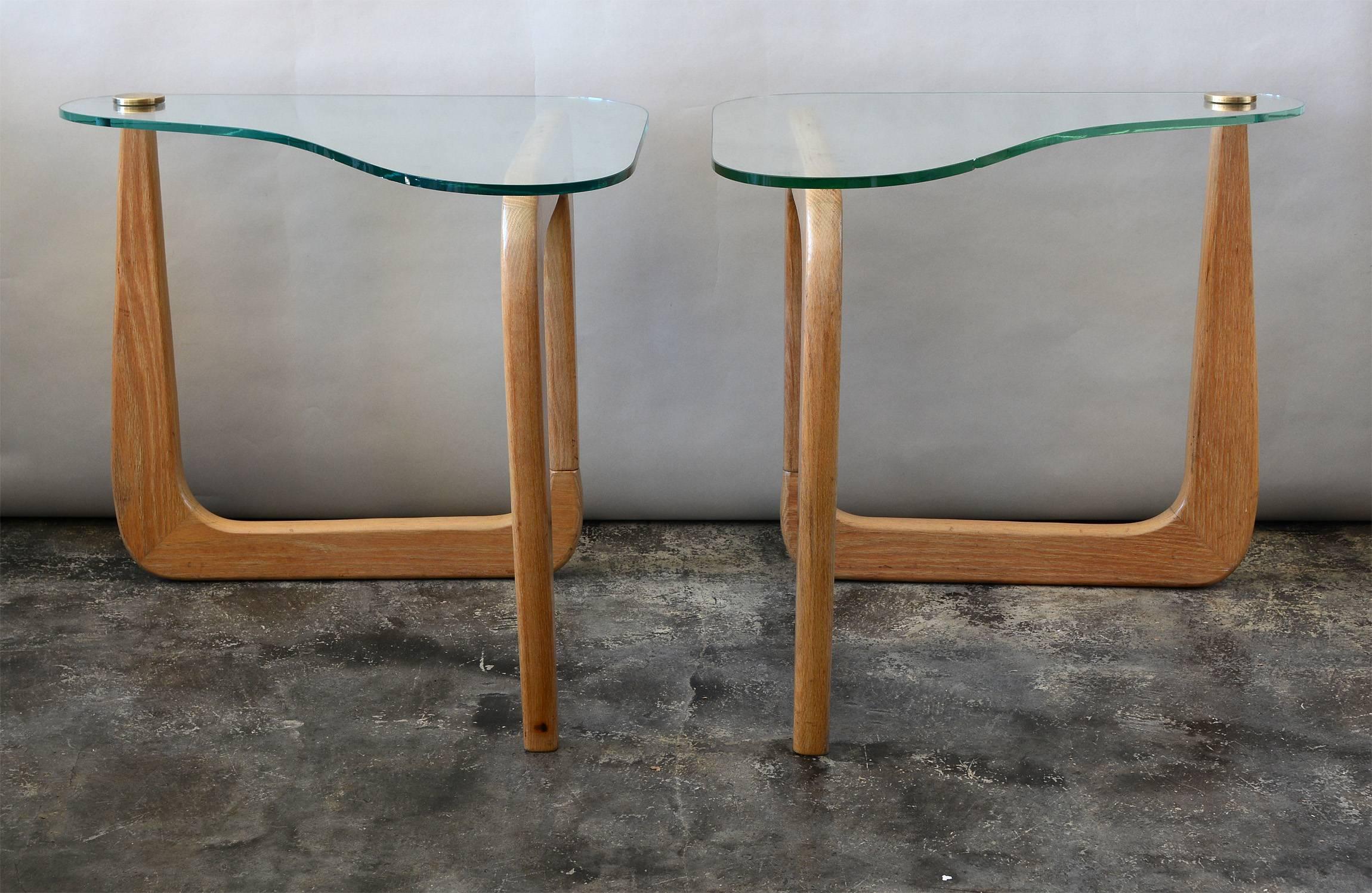 These biomorphic end tables have cerused oak bases and glass tops. A large brass disk holds the glass to one leg. There is some wear and discoloration in a few spots to the original finish. One top is the original pale green glass. There are two