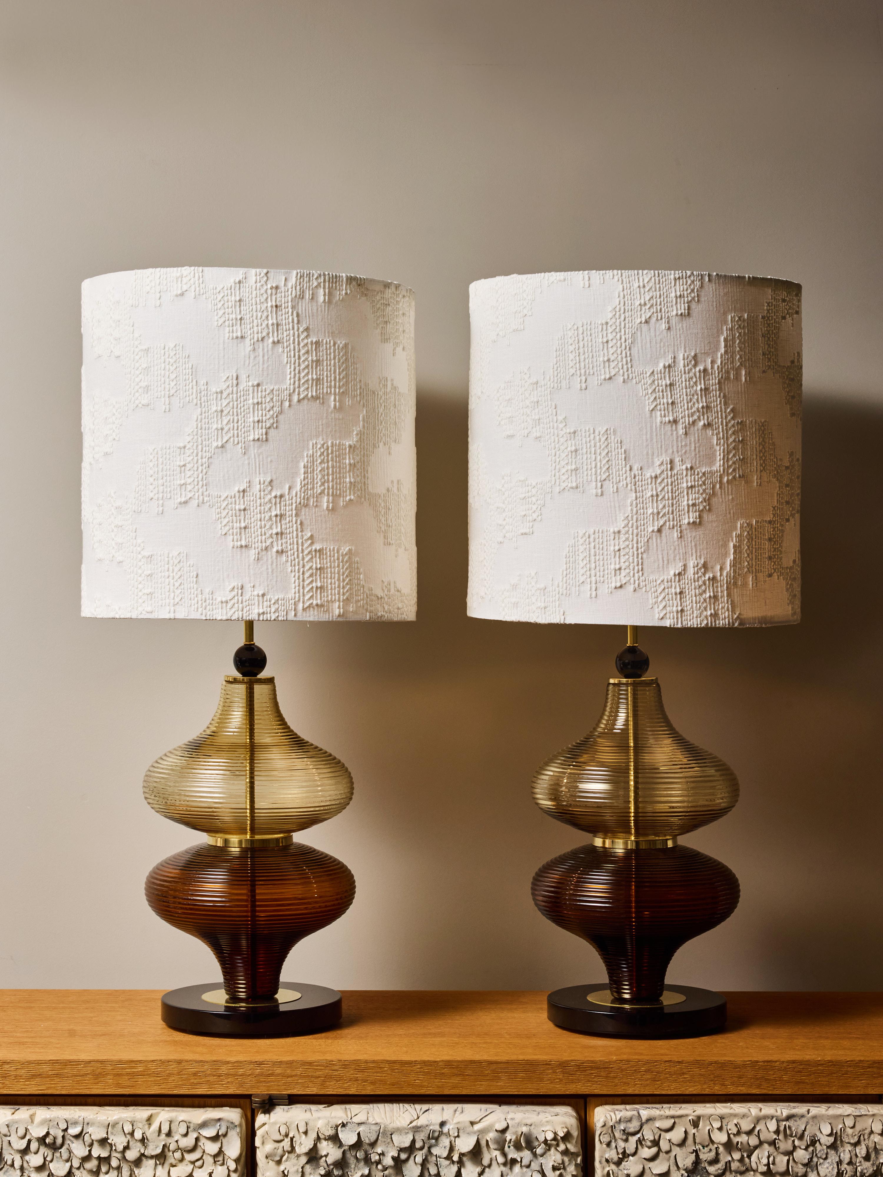 Questa elegante coppia di lampade da tavolo contemporanee è caratterizzata da basi scultoree in vetro formate a mano, composte da due livelli impilati e nervati nei toni graduati dell'ambra calda e del bronzo fumé. Le forme del vetro creano un gioco