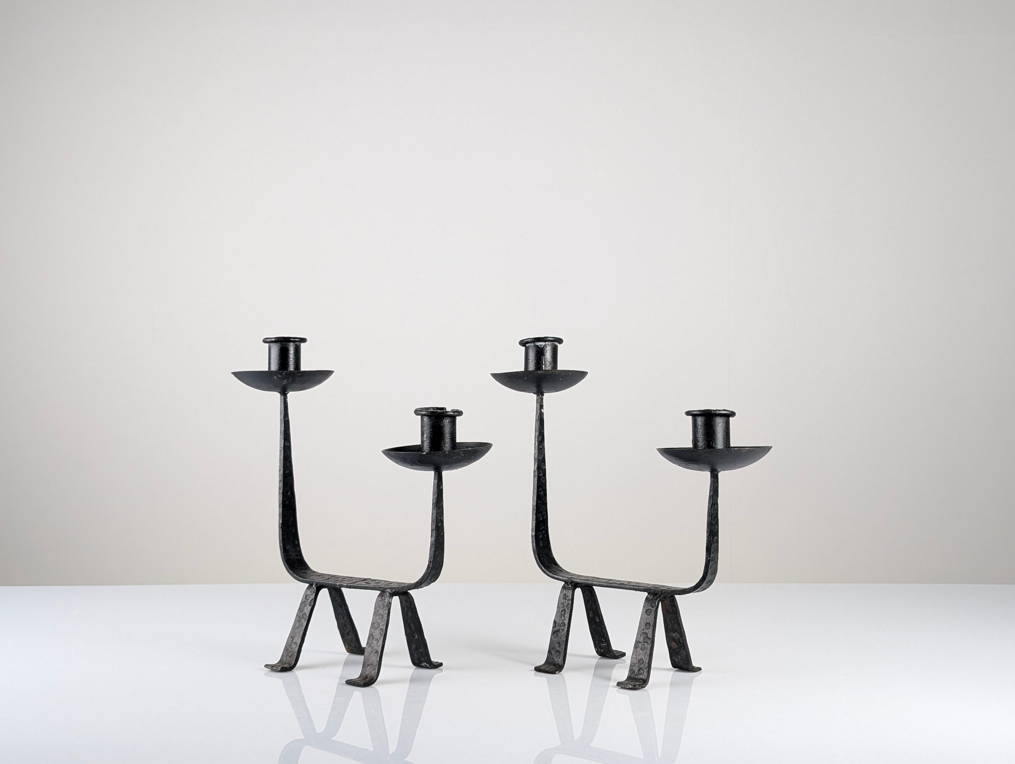 Pair of wrought iron candleholders made in Europe around 1950, featuring a sculptural design with clean lines and totemic presence. Each piece has two candle arms at different heights, creating a balanced and dynamic composition. The four-legged