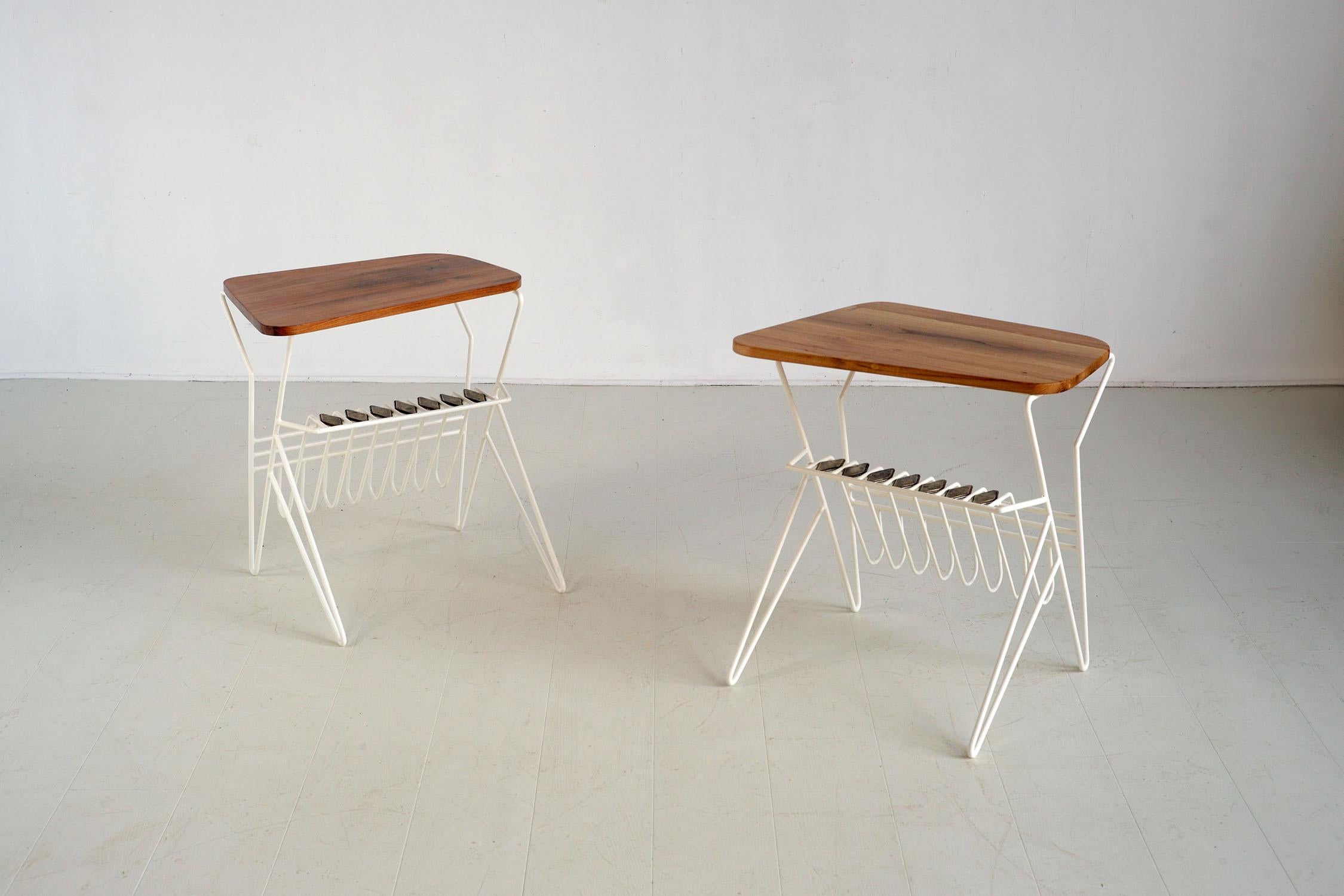 Elegante pareja de mesillas auxiliares de metal tubular lacado en blanco, Francia 1950. La perfilada base contiene un estante para periódicos y una repisa decorada con rombos de latón patinado. La parte superior de olmo macizo es asimétrica. El