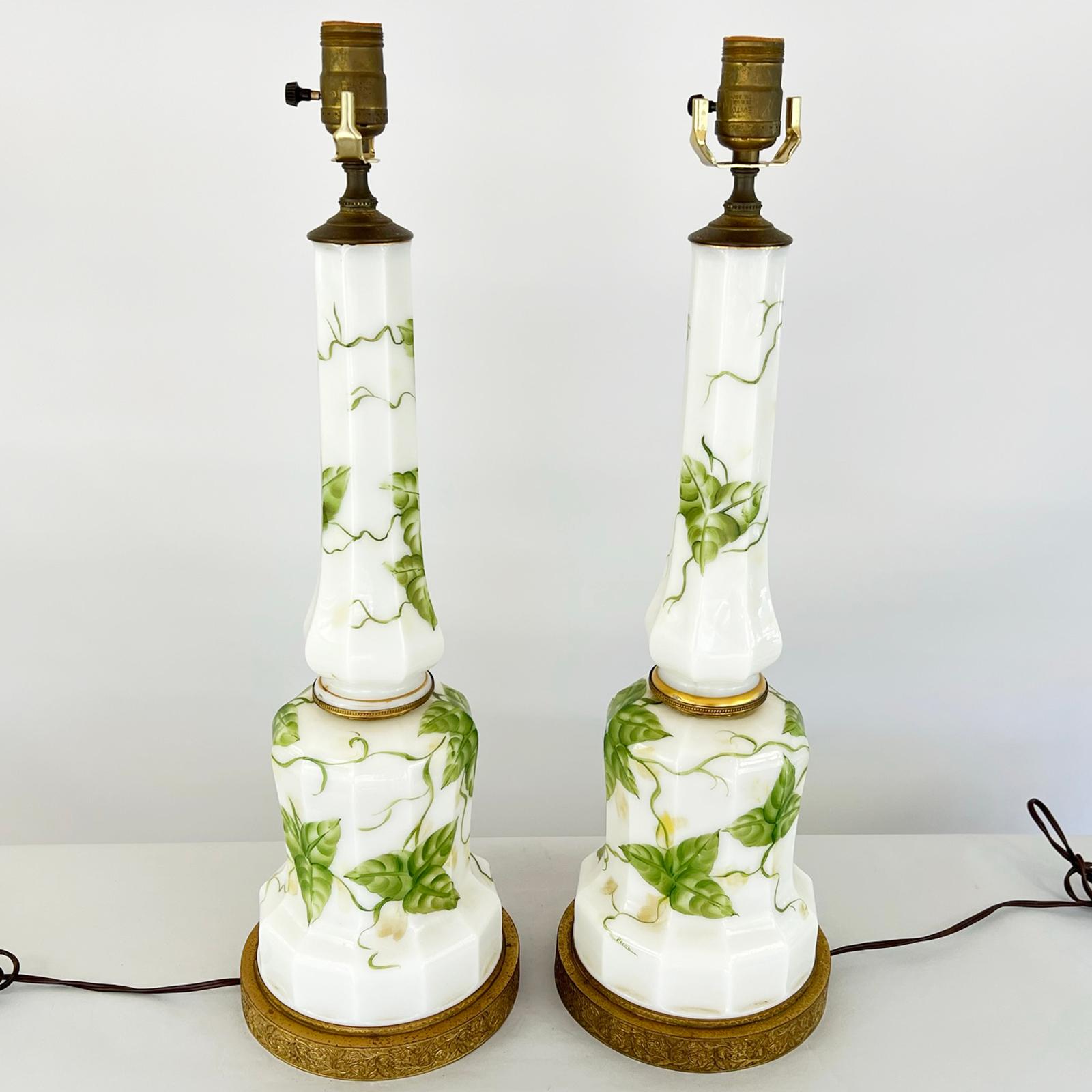 Pair of columnar table lamps, in Empire style, of milkglass. Each a round, faceted column, handpainted with scrolling vines of ivy. Lamped on round, pierced metal bases. Signed, 
