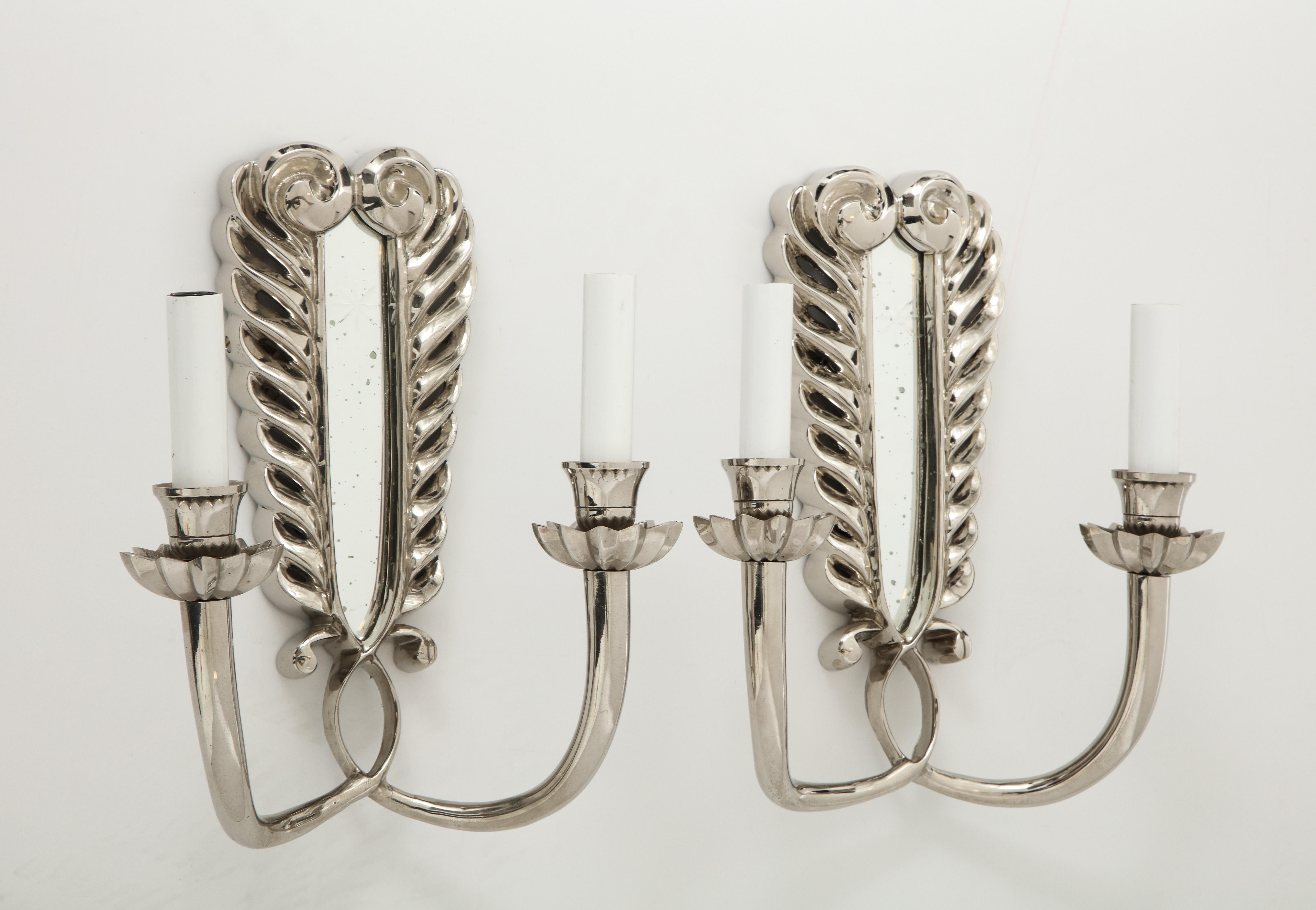 A pair of polished nickel double arm sconces with a mirrored back plate, surrounded by a scrolling design. The bobeche on each arm mimics a flower petal and adds an elegant touch to the frame. Priced at $2800 per pair.