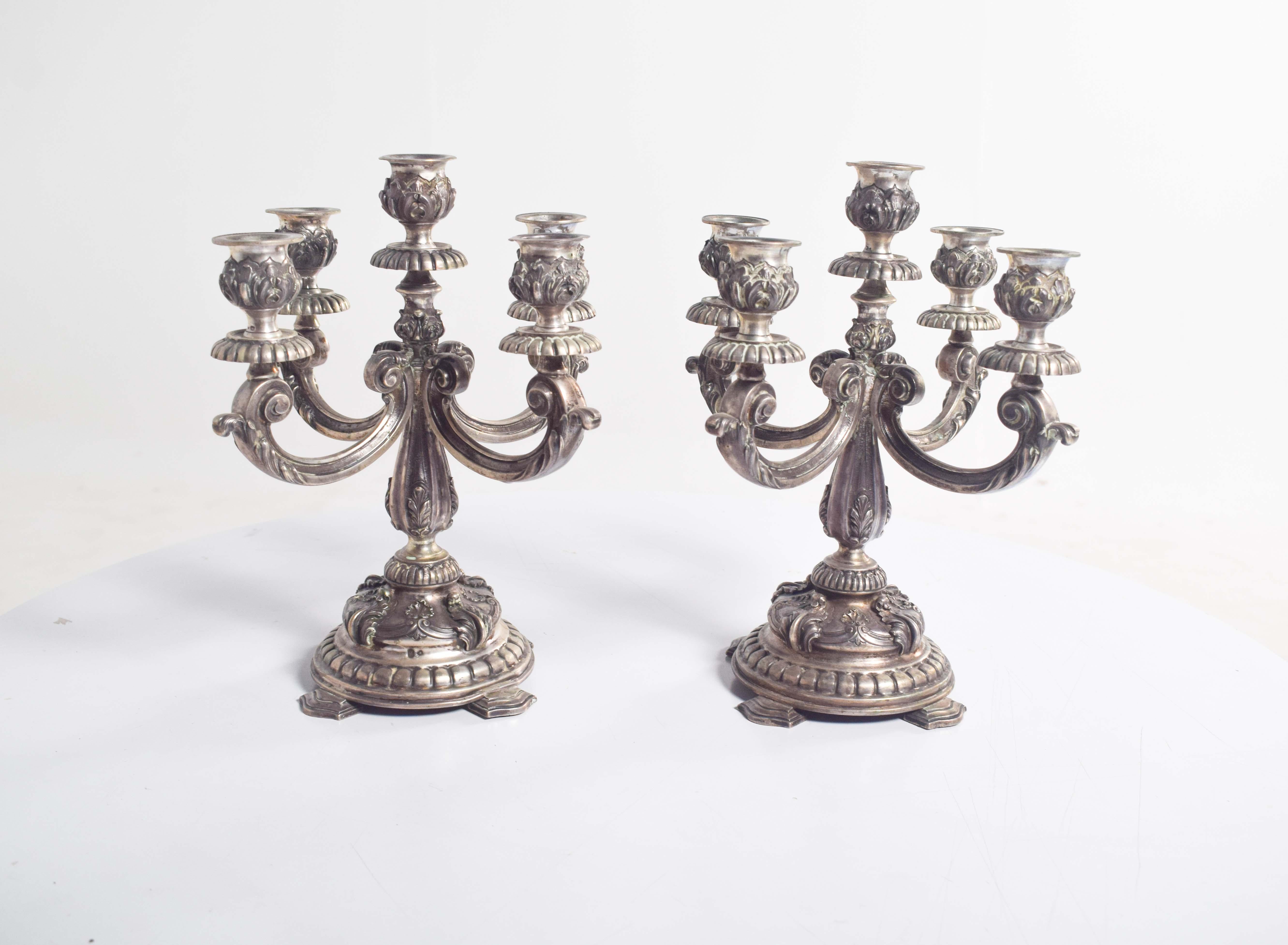 Questa splendida coppia di candelieri a cinque luci, realizzati in elegante argento 830, mostra un'armoniosa miscela di funzionalità e arte decorativa. Ogni candeliere poggia su una base rotonda a gradini sostenuta da quattro gambe robuste, che