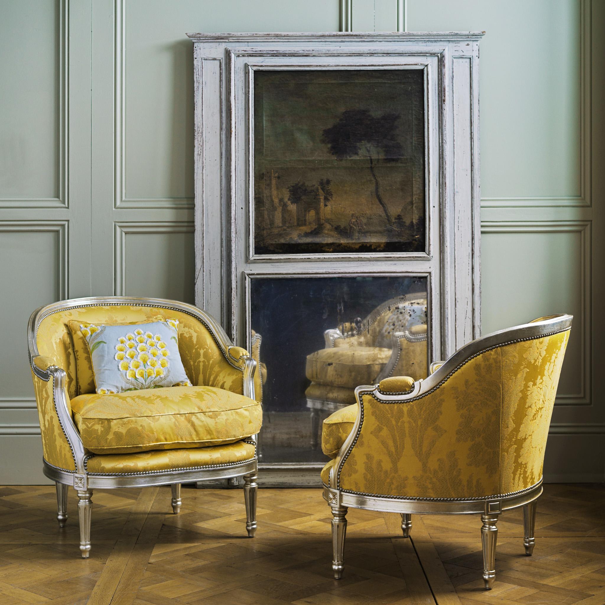 A pair of Hollywood Regency style marquise armchairs featuring elegant curves and a glamorous silver gilt wood finish. The chairs have a plump feather seat cushion for comfort and are upholstered in an Italian fabric by Rubelli with a blue scatter