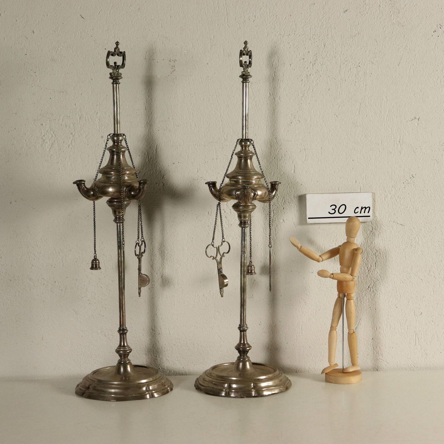 Pair of oil lamps made of molten shaped and chiseled silver. Brand of the silversmith at the bottom. Circular shaped base, shaped and ribbed shaft. The oil container has three spouts and a shaped handle with volutes. Three small chains hang through