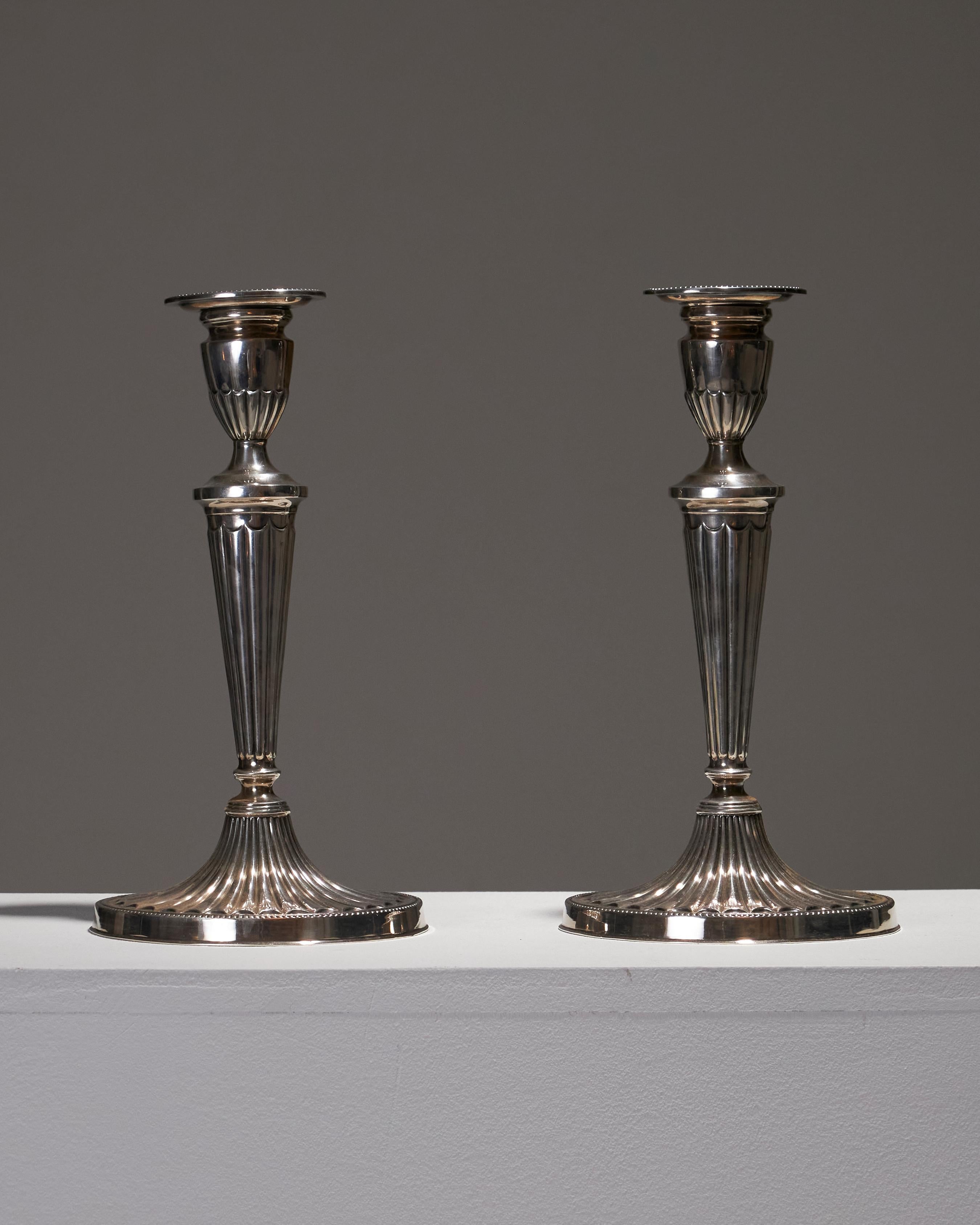 Coppia di candelieri argentati in stile neoclassico, realizzati in Inghilterra negli anni Venti. La loro silhouette sottile e scanalata poggia su una base svasata ornata da delicati dettagli di perline. Eleganti e senza tempo, questi candelieri