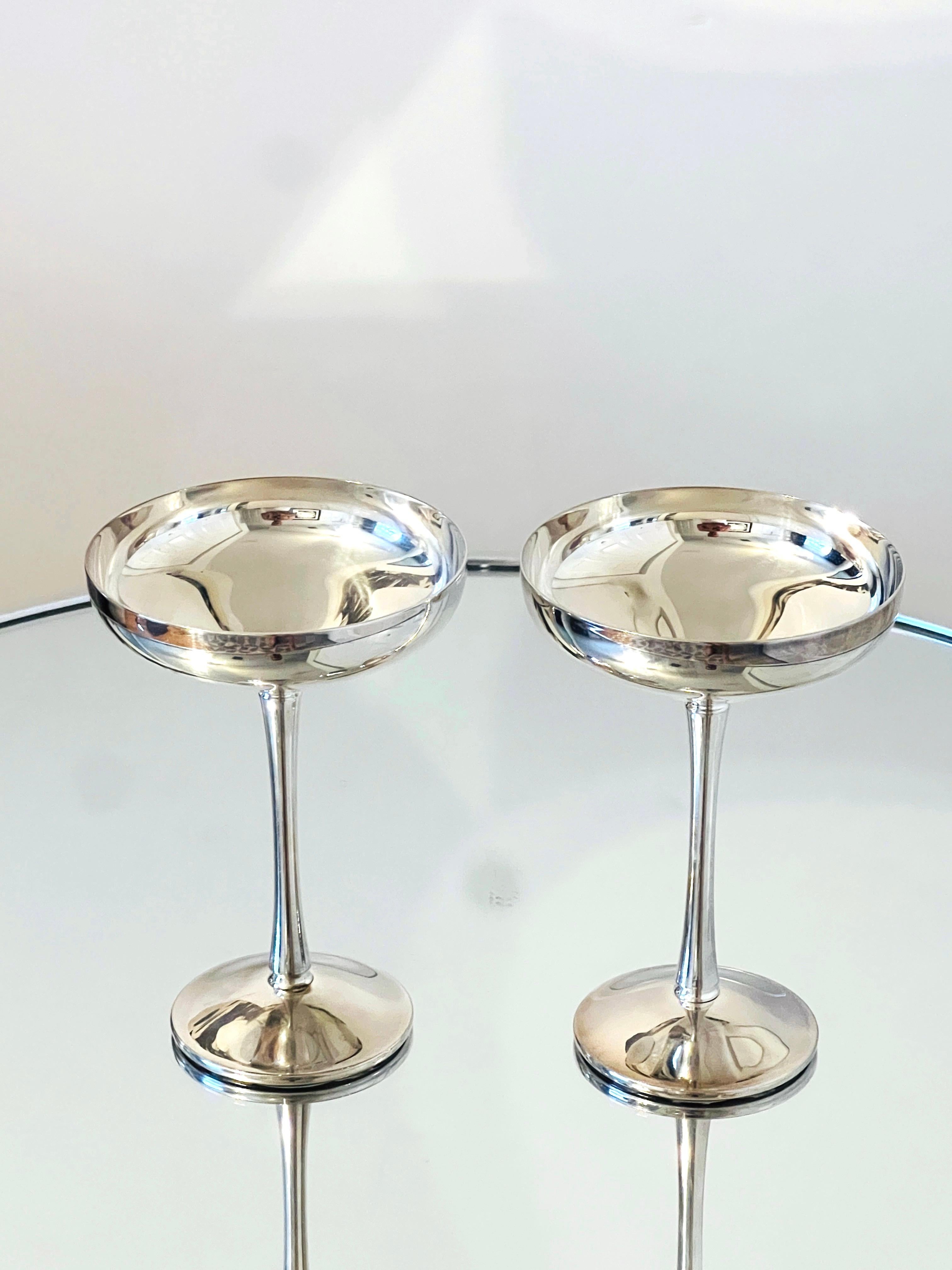 Pair of silver plated champagne coupes or toasting goblets by Kirk-Stieff. Feature elegant rounded saucers with tapered stems, and ring notches along the top and bottom of the stems. The coupes bare the company hallmark on the underside of each cup.