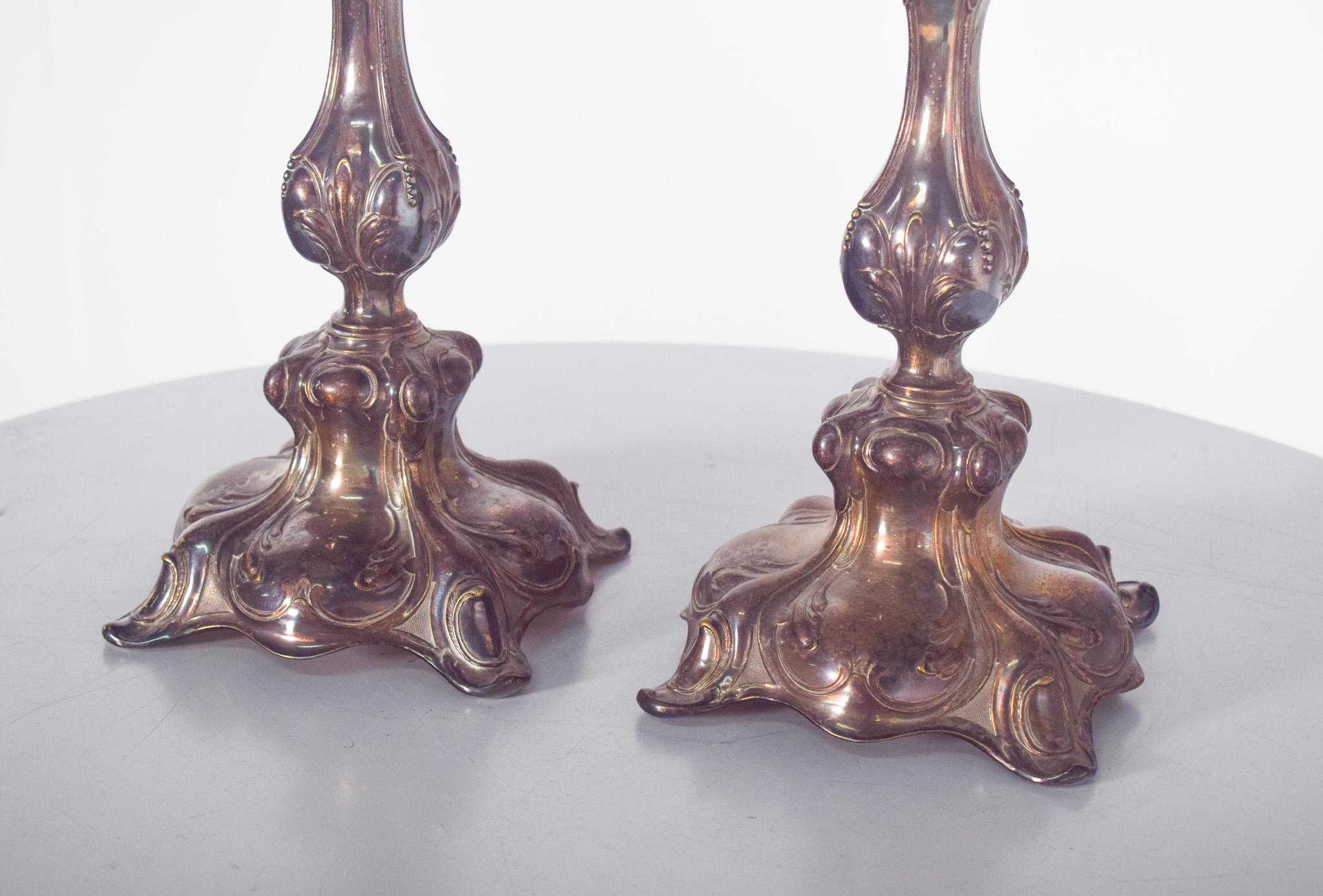 Coppia di candelieri in metallo argentato. Hanno una base ampia con quattro supporti arrotondati e una decorazione in rilievo con forme curve. Il corpo è stretto al centro e si allarga nuovamente nella parte superiore, con dettagli in rilievo che