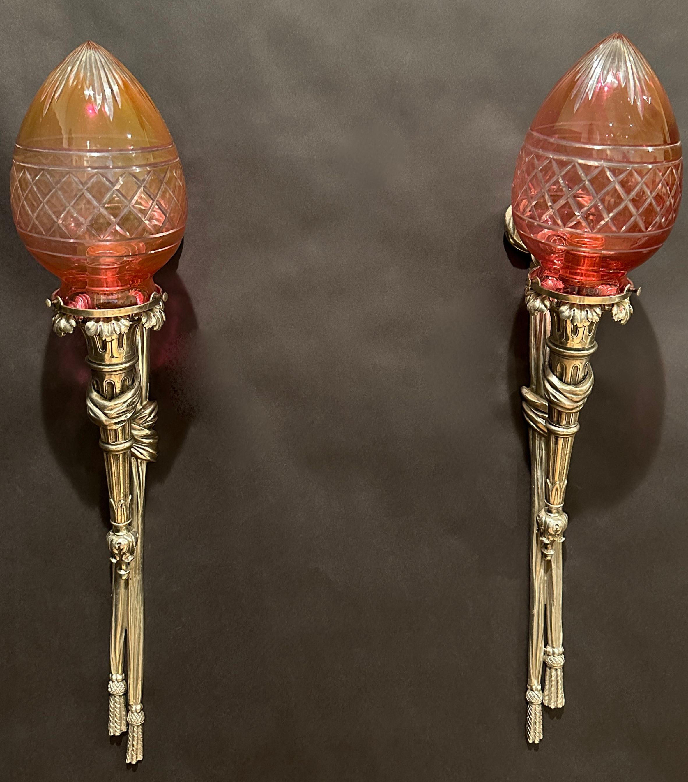 Pair of silvered bronze wall sconces in the Louis XVI style. Ruby cut to clear torpedo shaped glass shades. Sconce in the form of a ribbon with a bow knot a rosette and tassels and the torch held with a tied ribbon. 