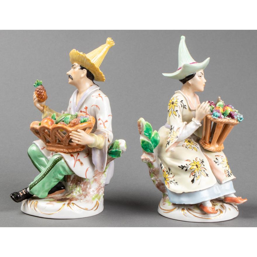 Pair of German Sitzendorf paint decorated porcelain figures, early 20th century, modeled as two seated fruit farmers in Chinoiserie attire, bases with blue crown over 'S' mark.