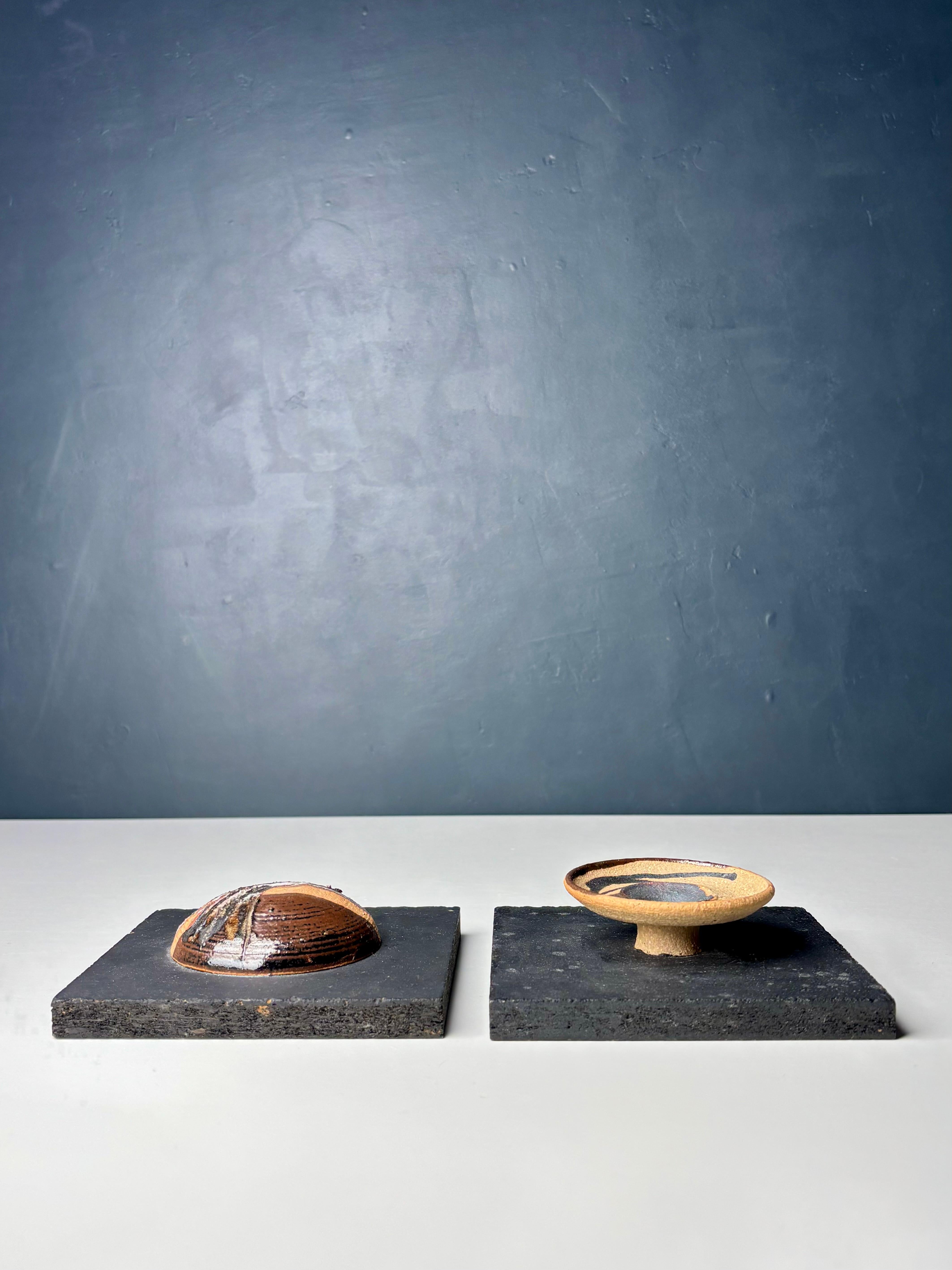 Pair of rustic Danish organic modern decorative earthcolored ceramic art wall plaques on square black mounts. Two different circular art pieces with shiny dark brown, gray and black colored glaze over the light brown raw material. Both mounts have a