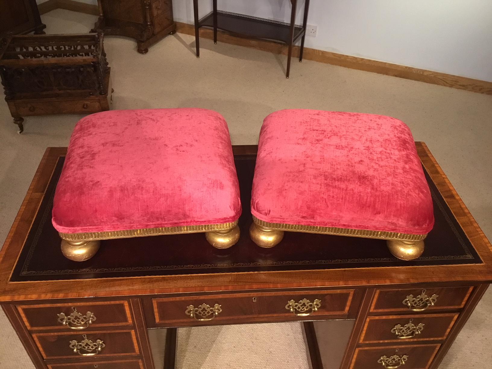 A pair of small square giltwood Victorian period foot stools. Each having a padded upholstered top newly re-upholstered in Clark and Clark majestic garnet velvet. Having a carved and gilded frieze and supported on four gilded bun feet with inset