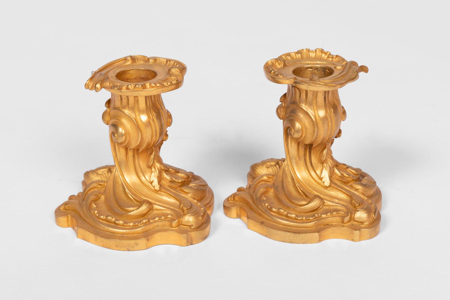 Pair of small rocaille style candlesticks in chiselled and gilt bronze standing on a scalloped base and top by a tumultuous shaft adorned with cartouches, acanthus leaves and stylized C-scrolls. Removable bobeche with curved edges also decorated