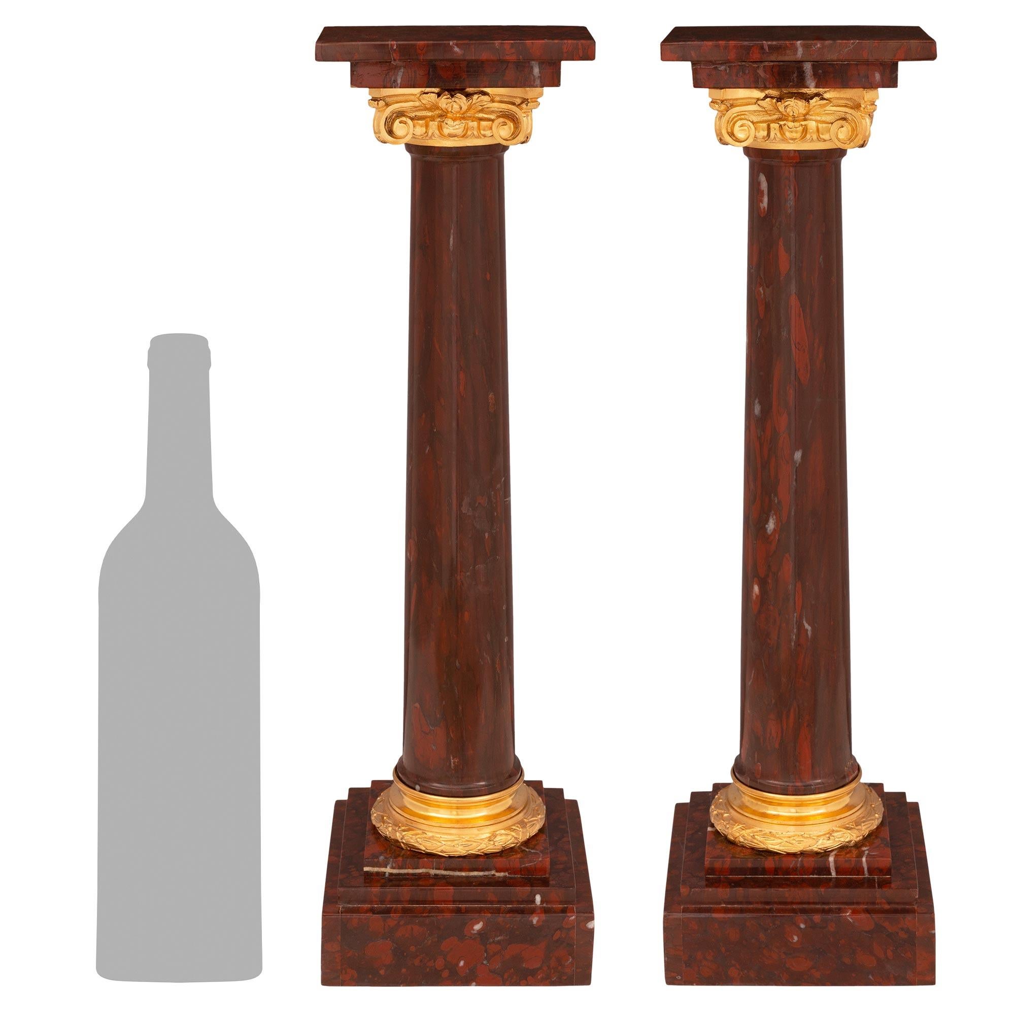 An elegant and most decorative pair of small scale French 19th century Louis XVI st. Rouge Griotte marble and ormolu columns. Each column is raised by a fine square base with stepped designs below circular mottled supports with richly chased tied
