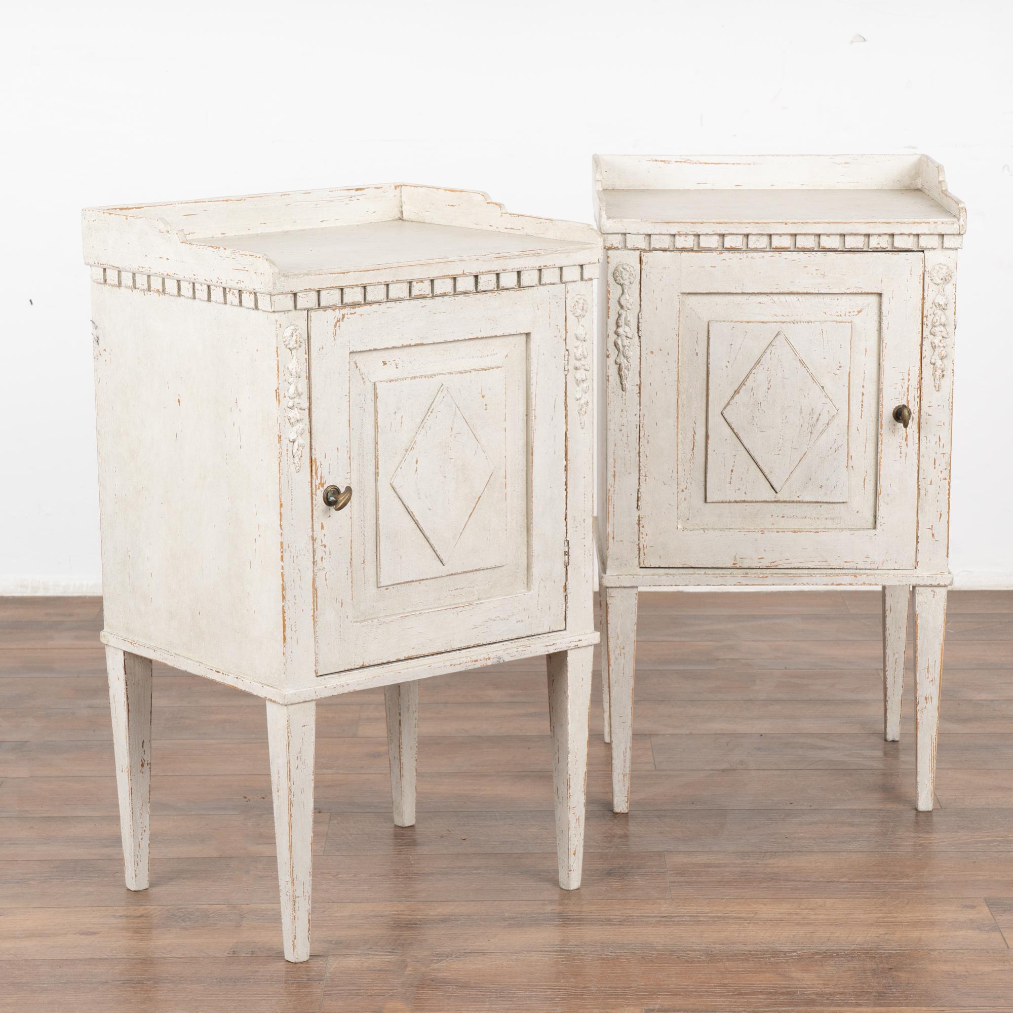 Pair, small Swedish country Gustavian white painted nightstands or side tables standing on tapered legs.
Applied floral carving accents the front along with dentil molding and diamond on panel door which opens with brass knob.
Professionally applied