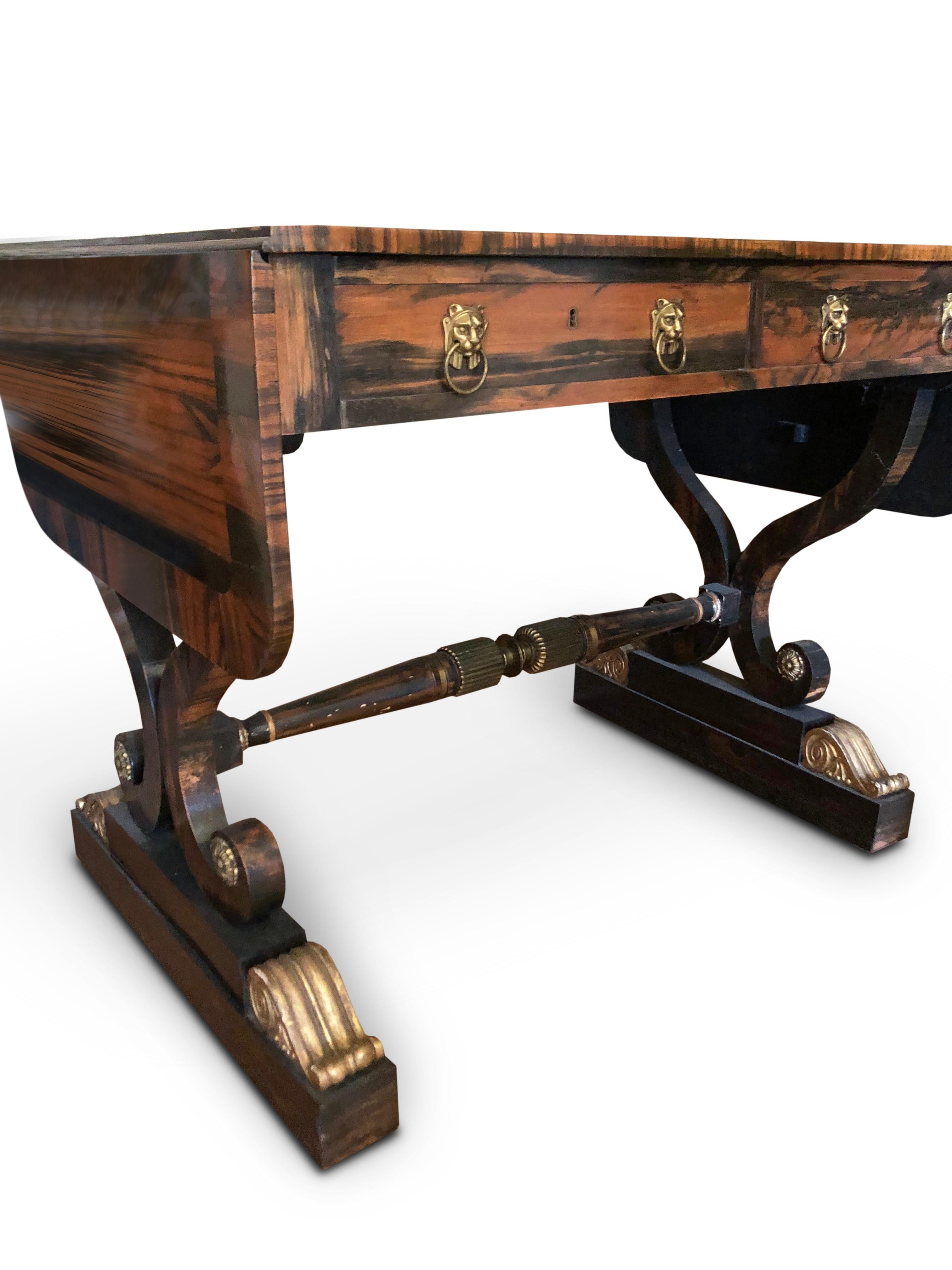 A very rare pair of George IV Parcel-Gilt  and Calamander Sofa Tables 
With rounded rectangular cross-banded tops, two frieze drawers with ring and lion mask handles, on scrolling pierced trestle supports joined by turned stretchers with ribbed