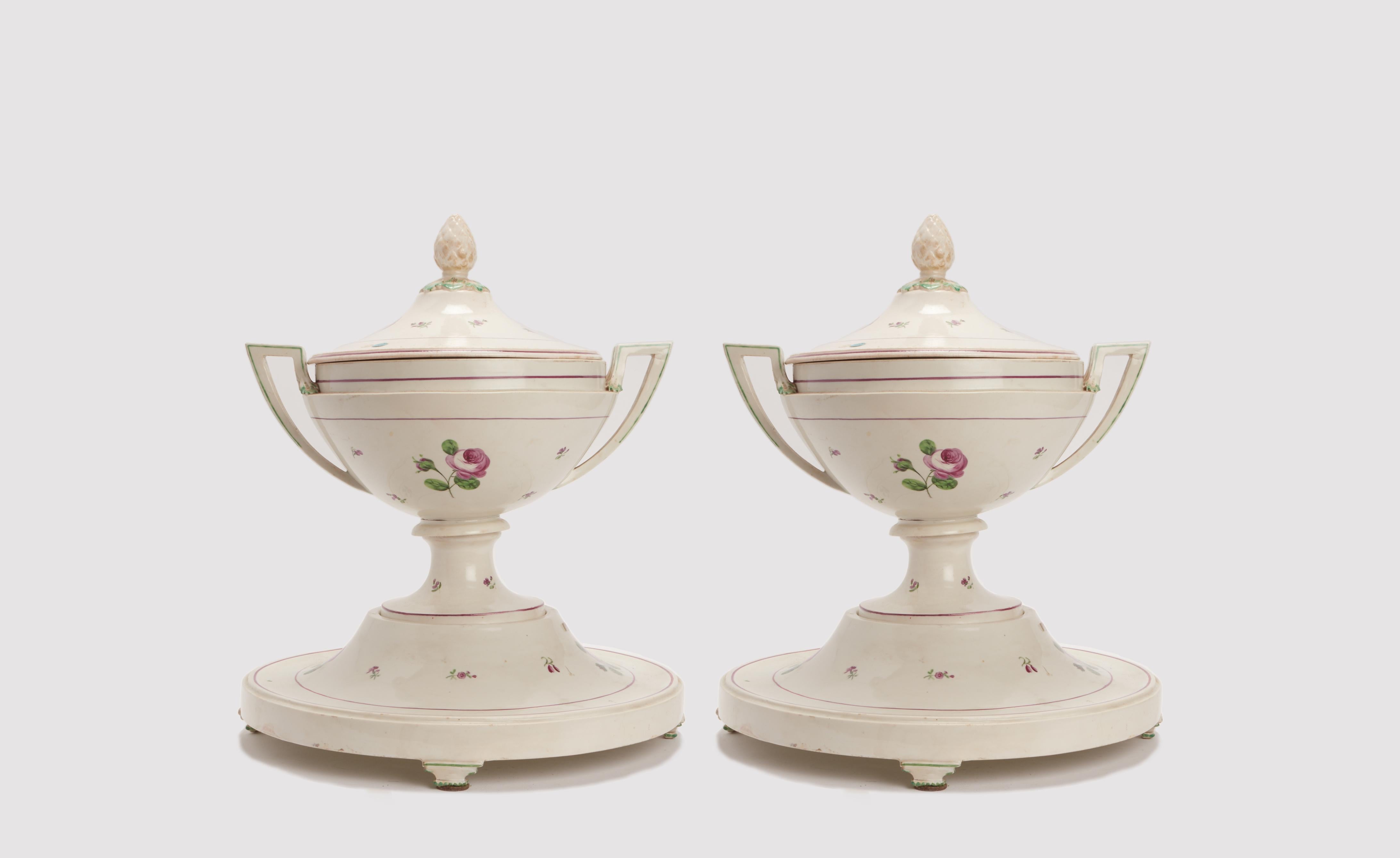 Coppia di zuppiere con base d'appoggio e manici angolati, decorate con motivi di rose su un campo bianco sottolineato da linee viola e verdi. Piedi distinti di forma geometrica, parte superiore del coperchio decorata con foglie e una pigna centrale.