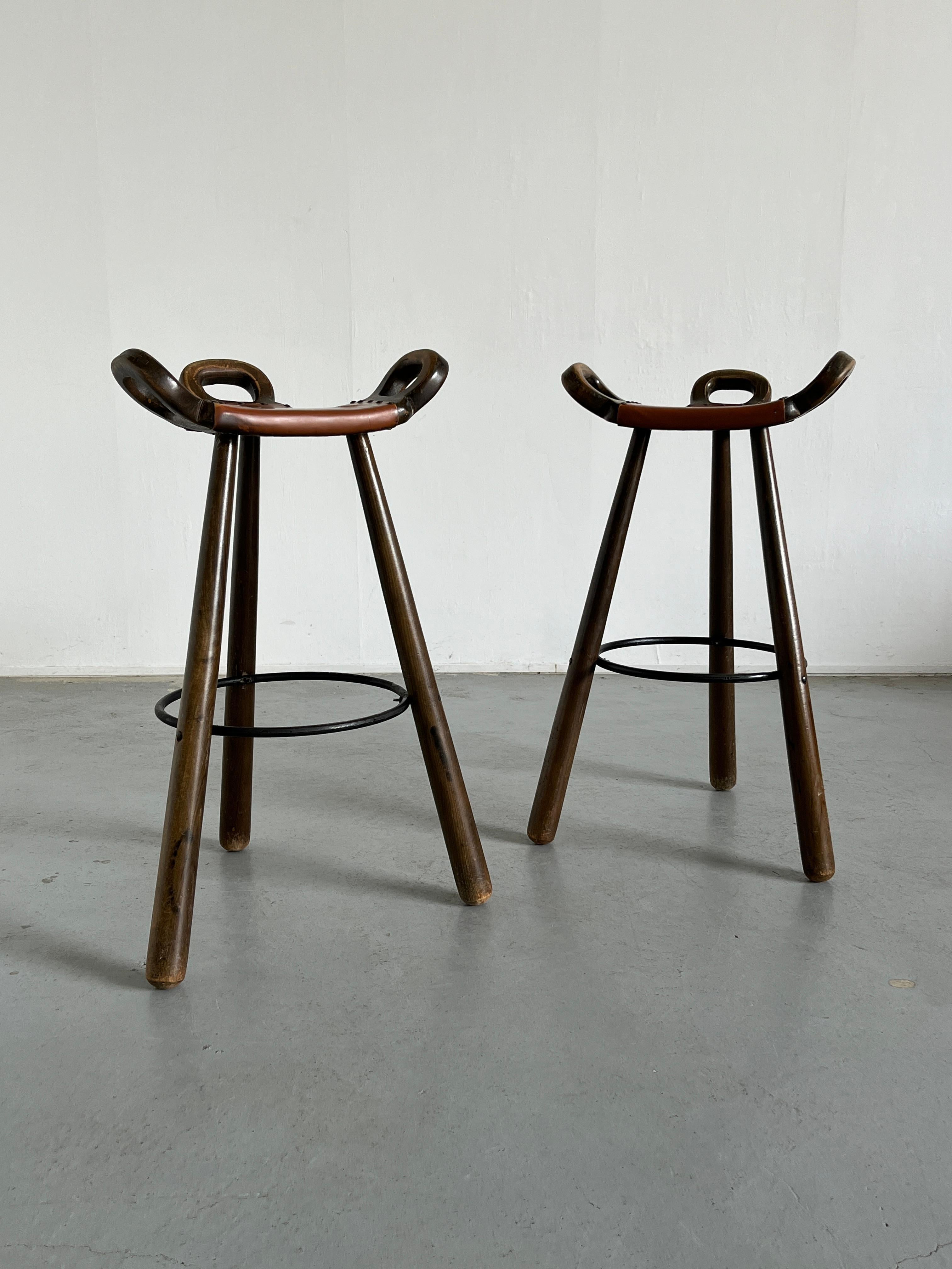 Two vintage “Marbella” bar stools attrributed to the Brazilian architect Sergio Rodrigues, unknown Spanish production of the 1970s. Three sturdy legs support the curved, saddle-like seat, striking shapes invoking Spanish tradition but also