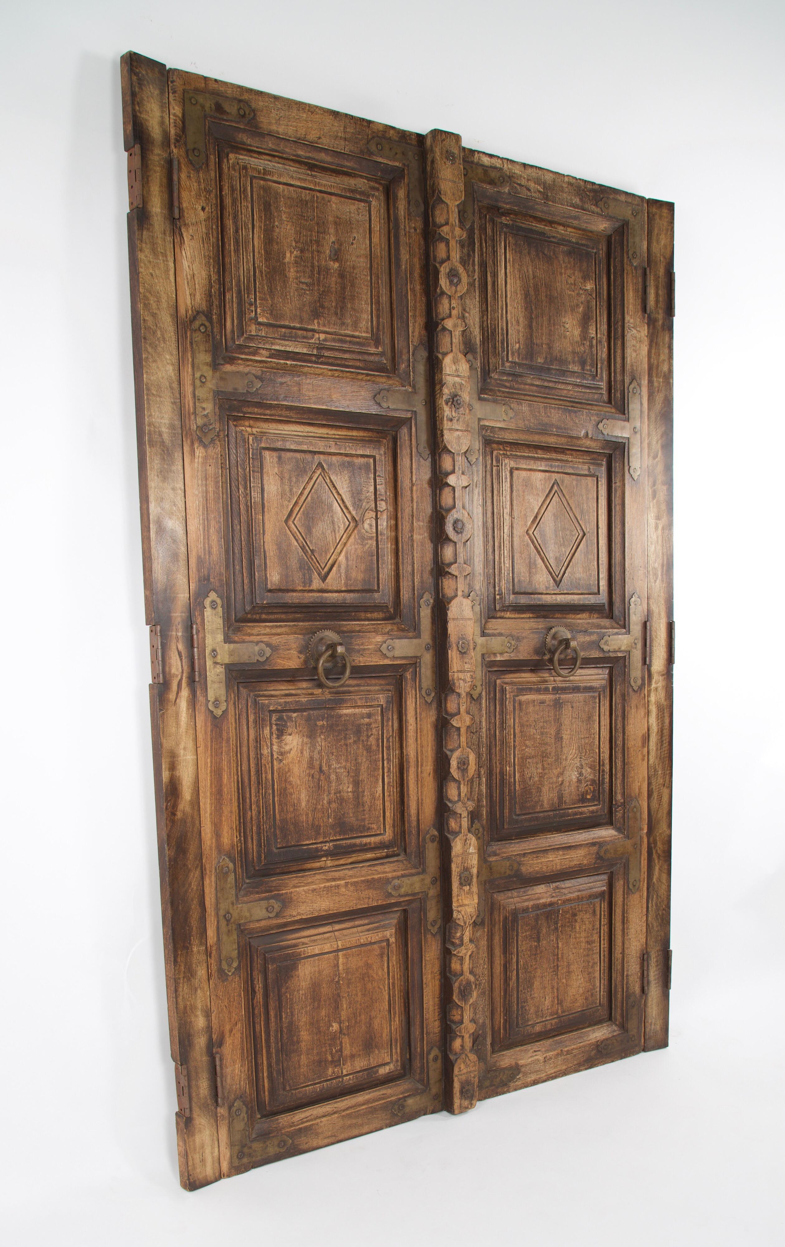 A pair of Spanish Colonial mesquite wood doors with brass door knockers also used as pulls to open the doors, brass straps and brass clavos. Four recessed panels on each door with a diamond pattern carved into one panel on each door. An elaborate