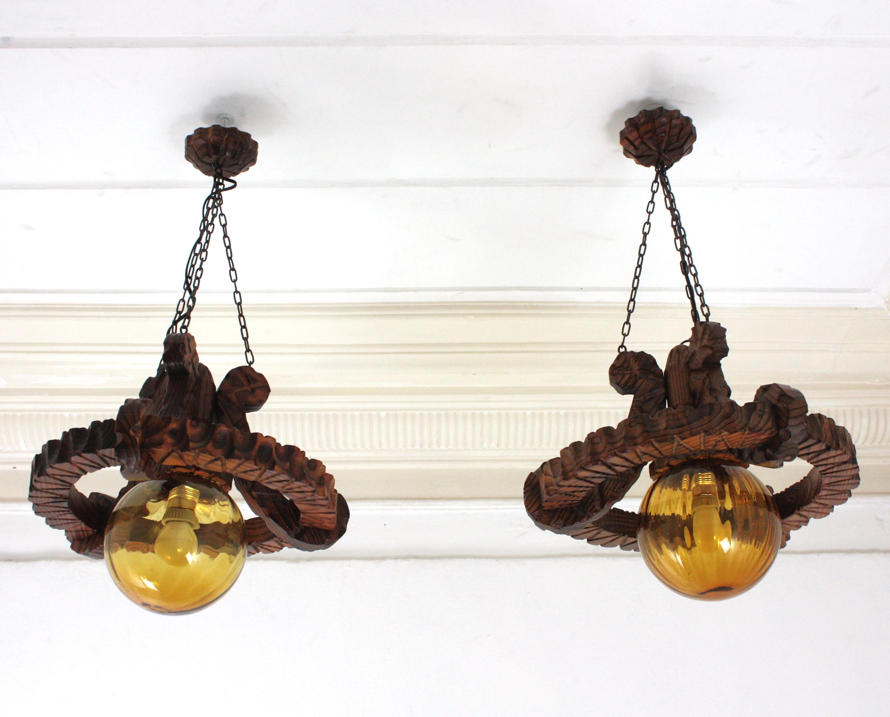 Pair of handcarved pine wood ring ceiling light fixtures with amber glass globe lampshades. Spain, 1940-1950.
This pair of ceiling light pendant lamps have an eye-catching construction. The ring backplates in carved wood hold an amber glass globe