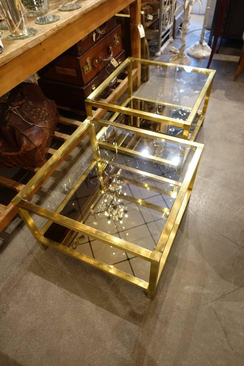 A gorgeous pair of elegant French quadratic brass tables. With smoked mirror glass shelf below and clear glass above. These tables can be used as both lamp and side tables, but also coffee tables. Stunning items.