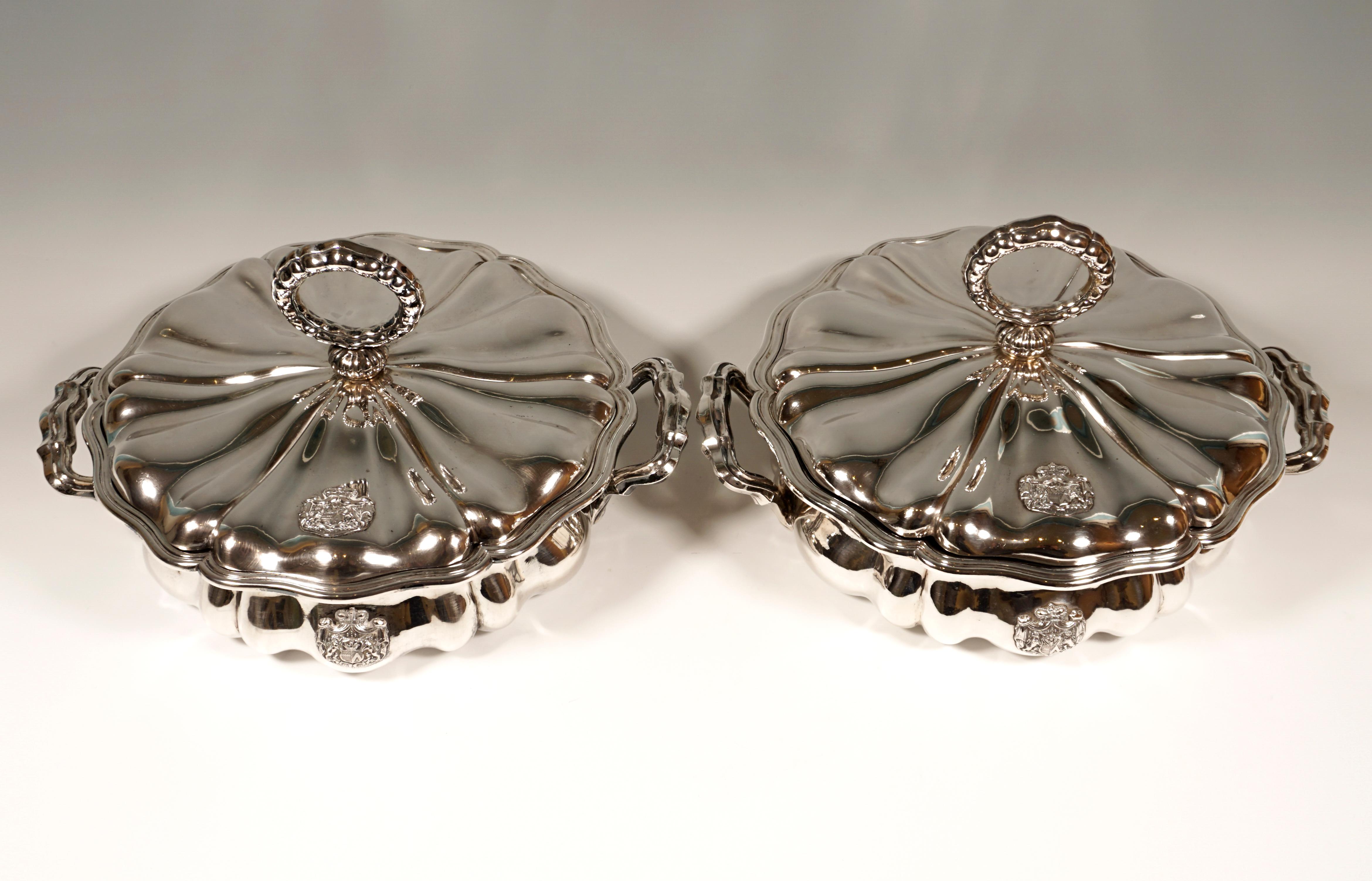 Two silver lidded terrines with a metal insert in a baroque style. 
Round basic shape, slightly bulbous body with incorporated ridges and curved areas. Ornate handles are attached to both sides. The top edge of both the bowl and the lid, as well as