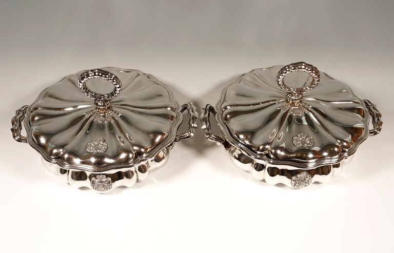 Two silver lidded terrines with a metal insert in a baroque style. 
Round basic shape, slightly bulbous body with incorporated ridges and curved areas. Ornate handles are attached to both sides. The top edge of both the bowl and the lid, as well as