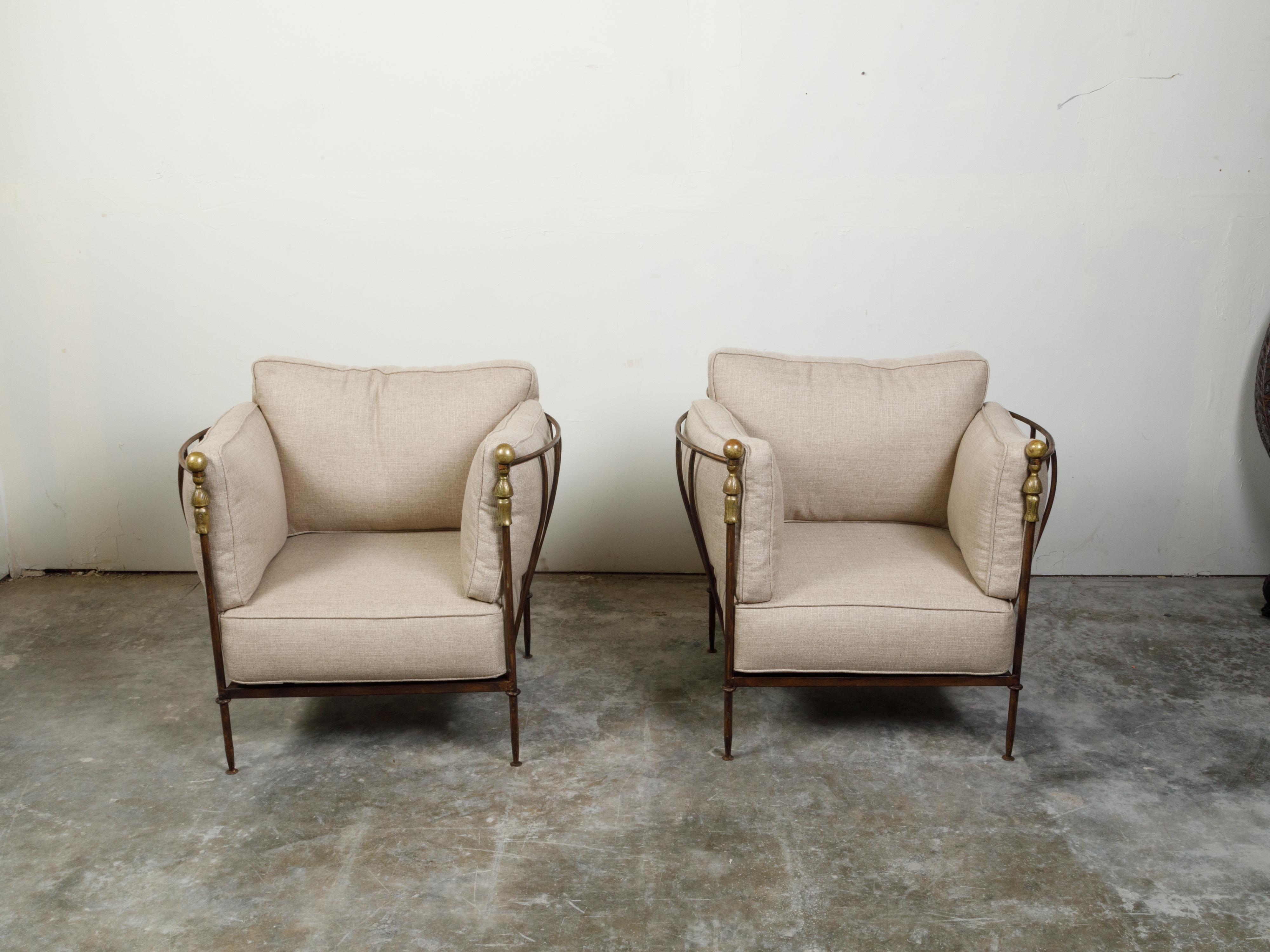 A pair of iron and brass club chairs from the mid 20th century possibly by Michael Taylor, with linen upholstery. Created during the midcentury period, each of this pair of club chairs features an iron structure with curving arm supports, accented