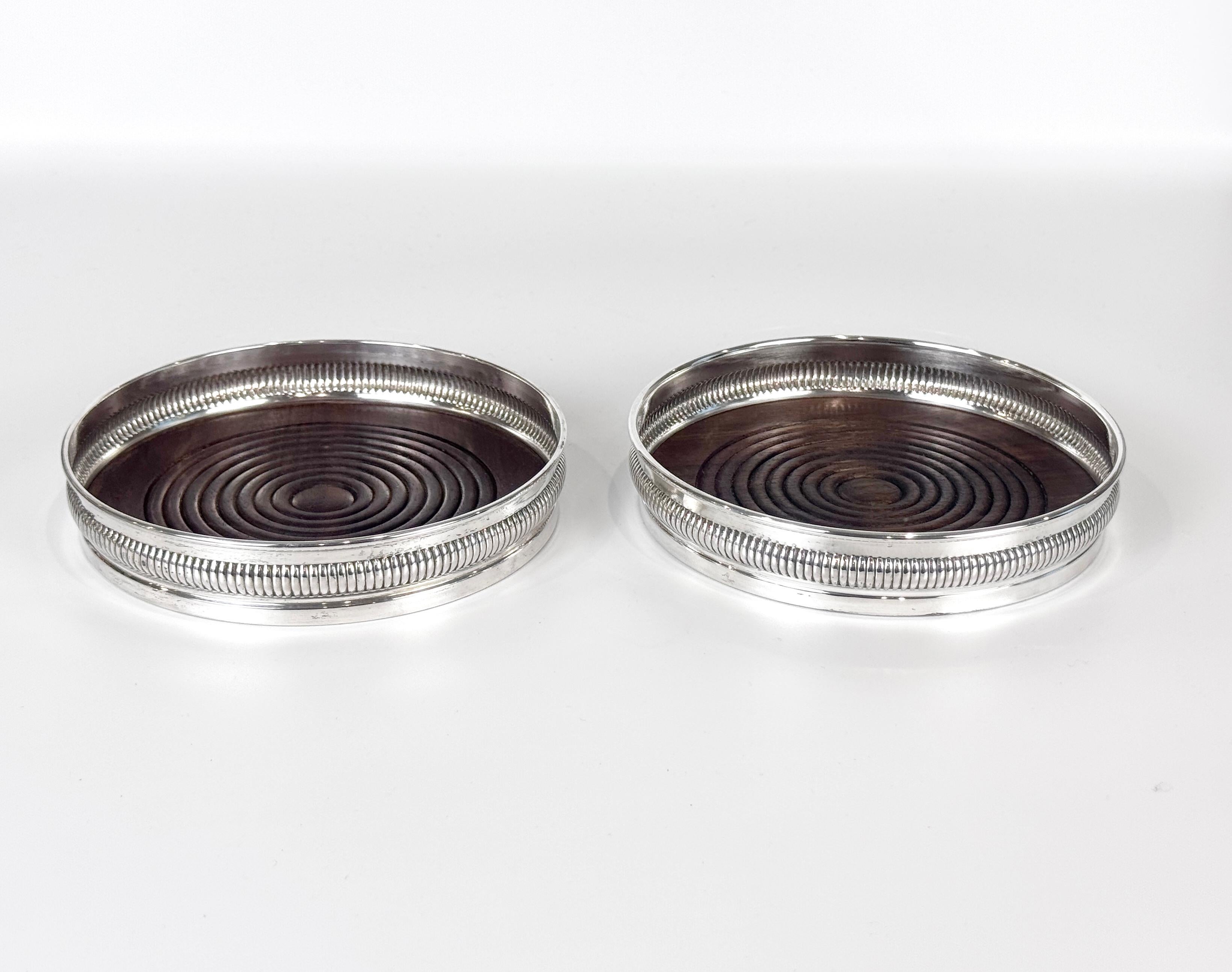 Pareja de posavasos de plata de ley y madera torneada, cada uno con la marca Sterling en la base. Rodeados por un borde estriado, estos posavasos tienen una sencillez chic que encajaría tanto en una mesa tradicional como en una más moderna. A la vez