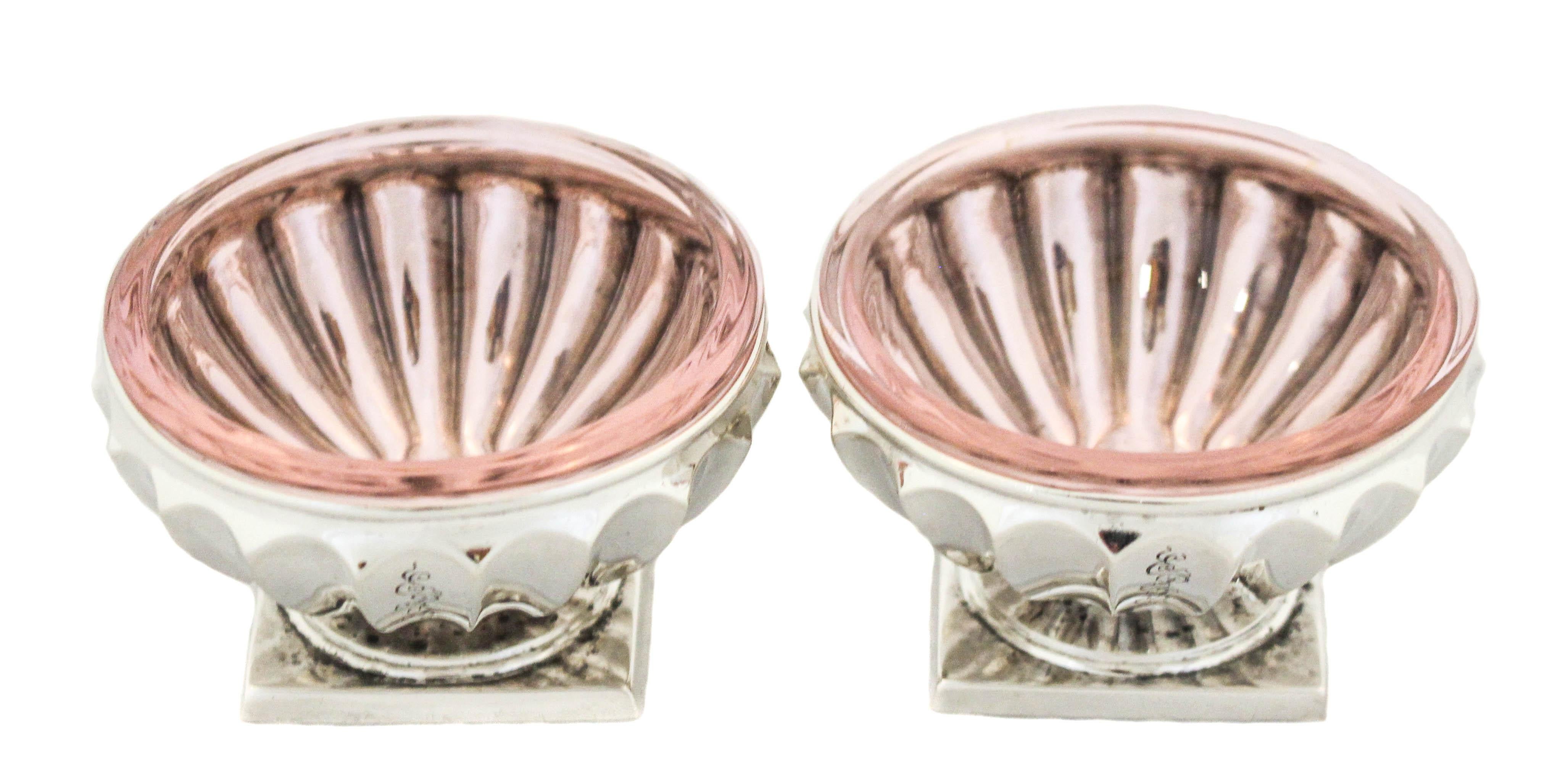 Being offered is a pair of sterling silver salt cellars by the world renowned Tiffany & Company. They have a square shaped base with a ribbed body. They each have a light purple glass liner that fits in perfectly to protect the silver from the salt.
