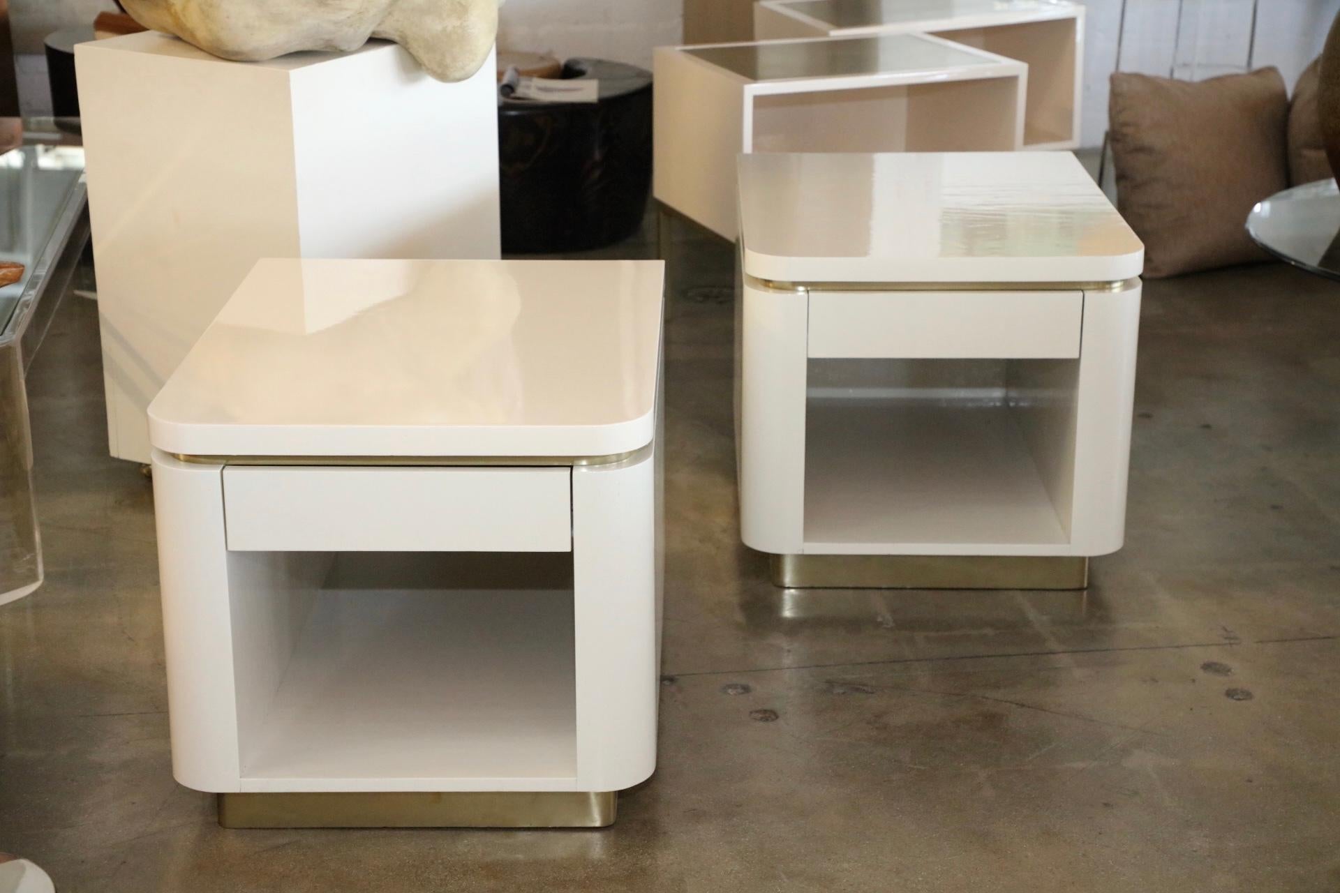 A pretty pair of Steve Chase designed nightstands or end tables. They are finished in ivory lacquer and brass. The brass is wrapped. There is an imperfection o the back corner, see photo. There are some imperfections as well as handling marks.