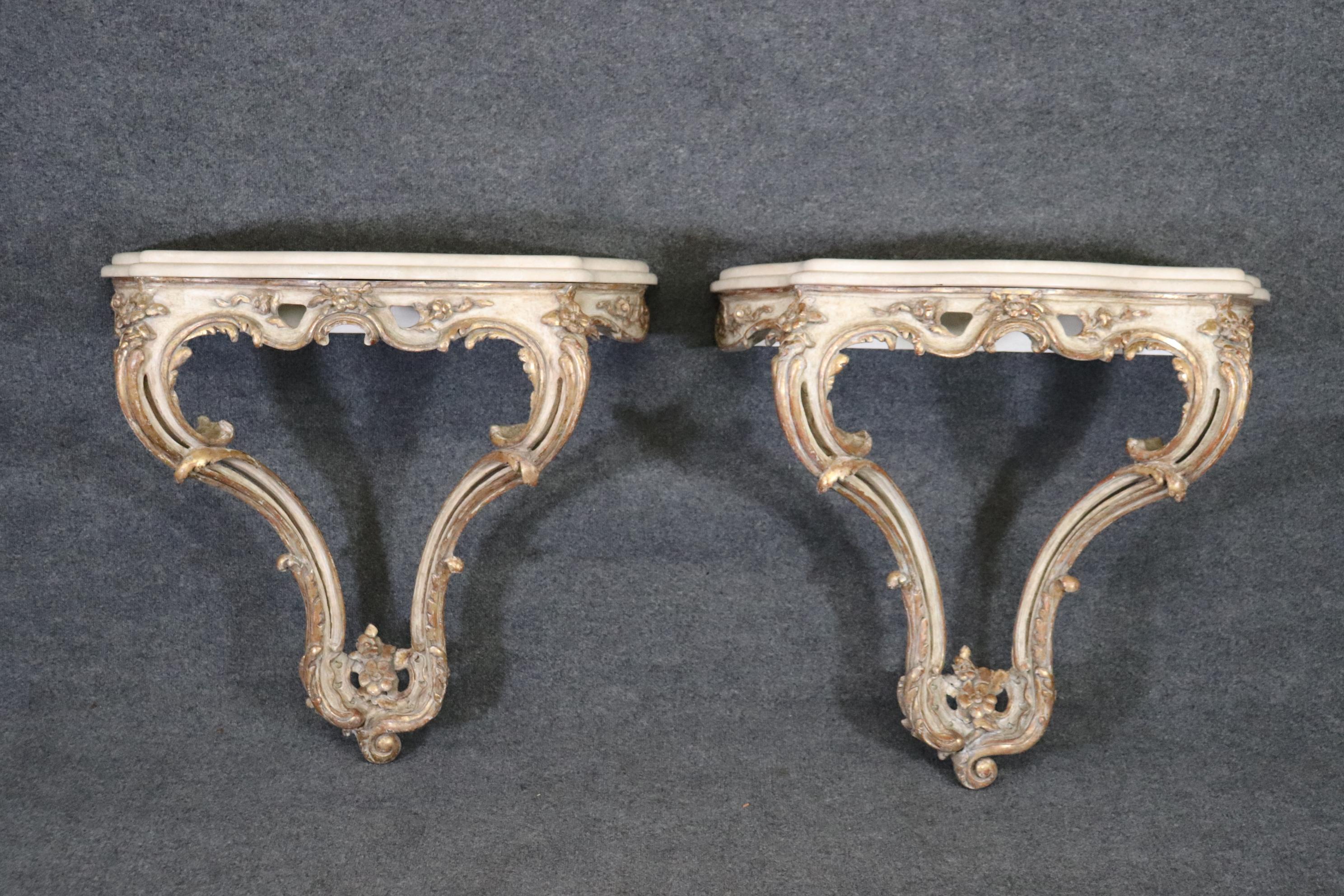 This is a gorgeous pair of wall-mounted Louis XV style painted and gilded consoles. They are painted in an antique white color, a cereamy color and gilded with gold. The tops are some form stone, perhaps marble or travertine. The tables measure 35