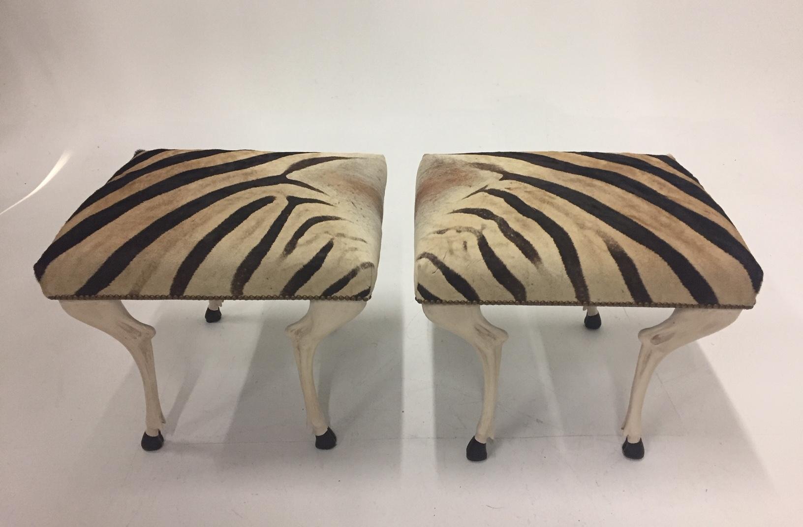 Wonderfully designed custom zebra ottomans having sculptural elegant painted wood legs that take their inspiration from a graceful hoofed animal, expertly upholstered in authentic zebra hide and finished with French hobnails. Color palette is cream