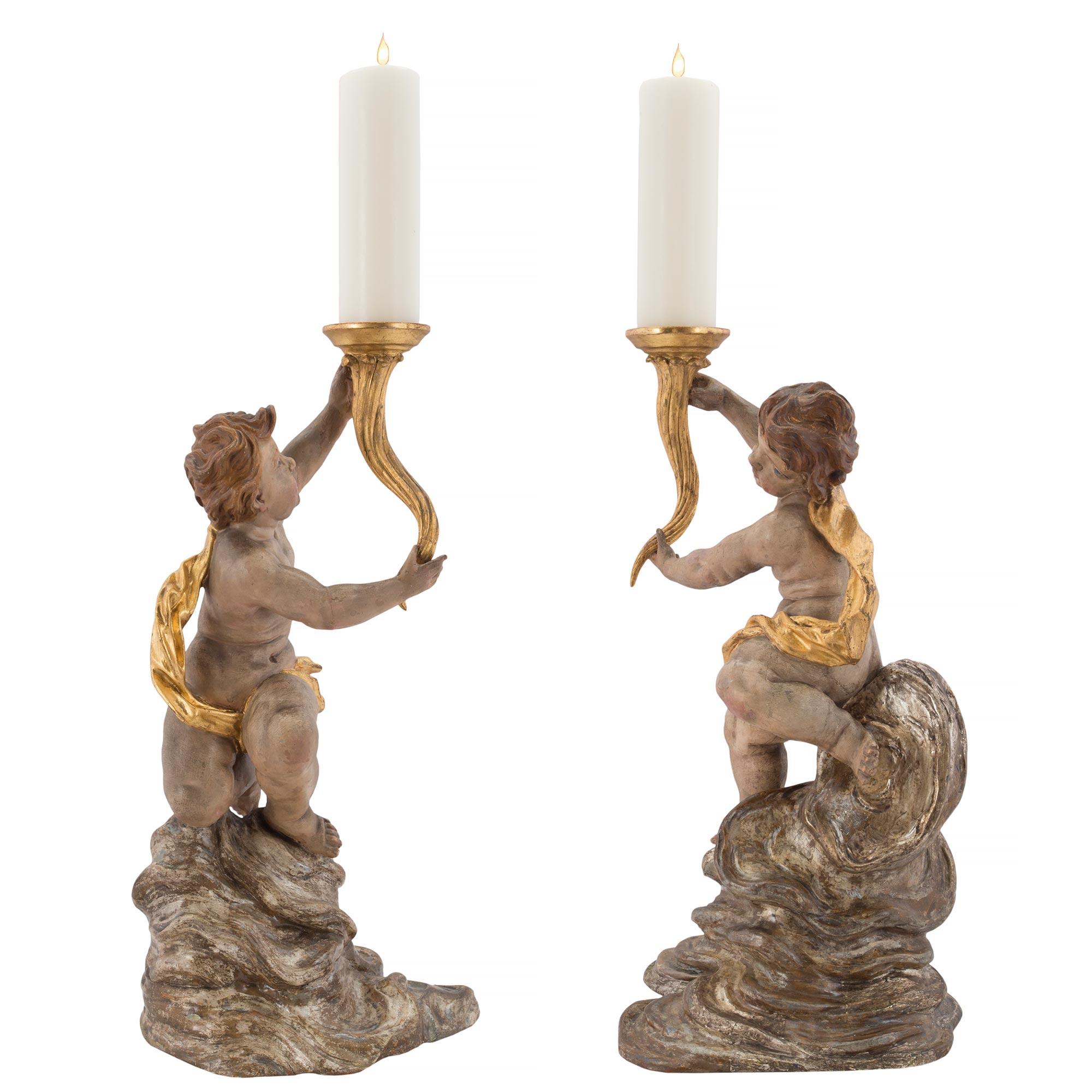A pair of stunning Italian early 18th century candle stands from northern Italy. Each charming cherub rests on polychrome silver leaf designed clouds. The patinated cherubs are adorned with a flowing giltwood drape and raise up a cornucopia styled