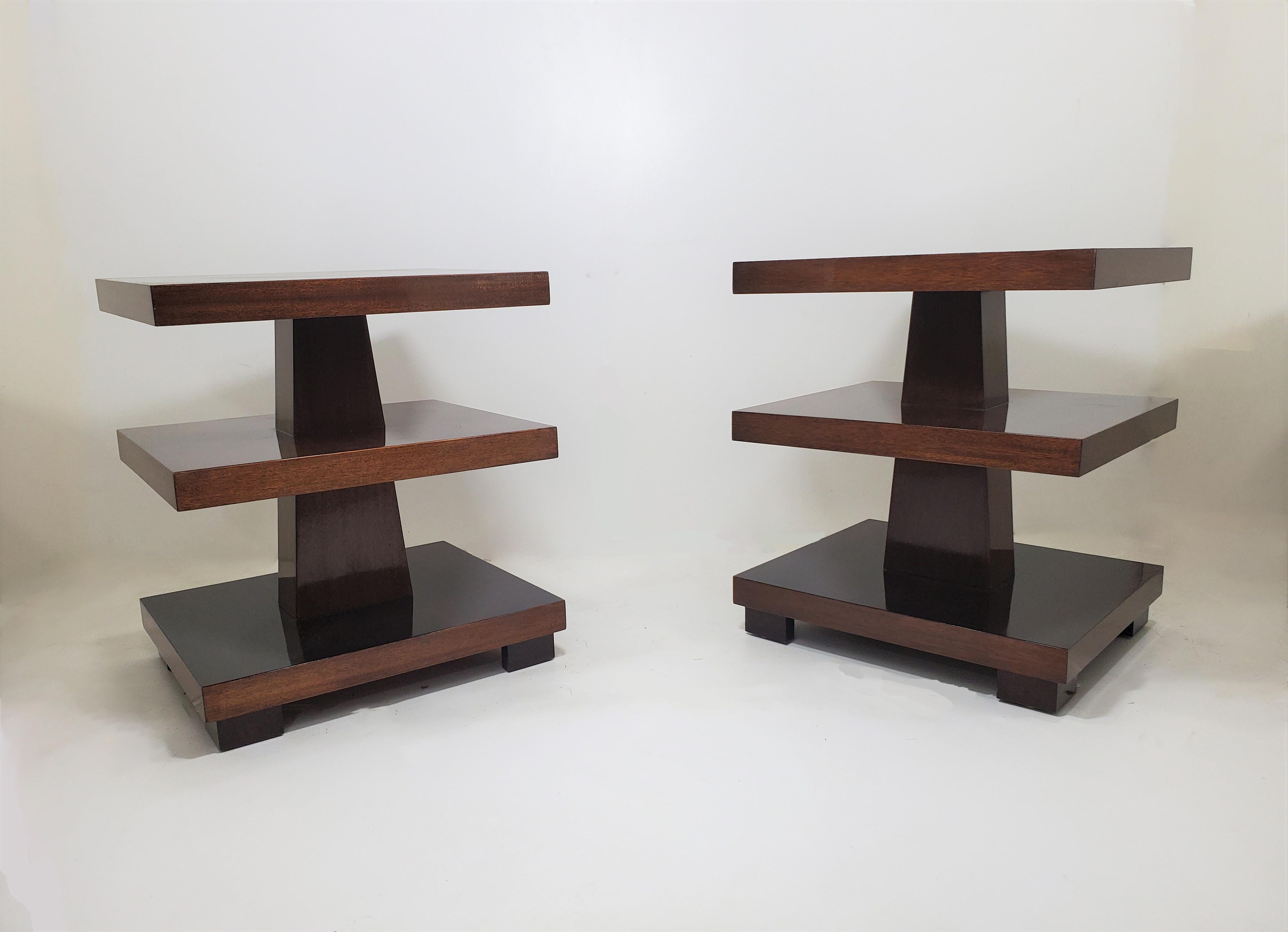 A pair of elegant and imposing highly sculptural, Art Deco rectangular three tiered tables of cubist design in a deep cognac /dark amber colored mahogany with central shaft dissecting the shelves and the whole raised on cube feet.
The angular form
