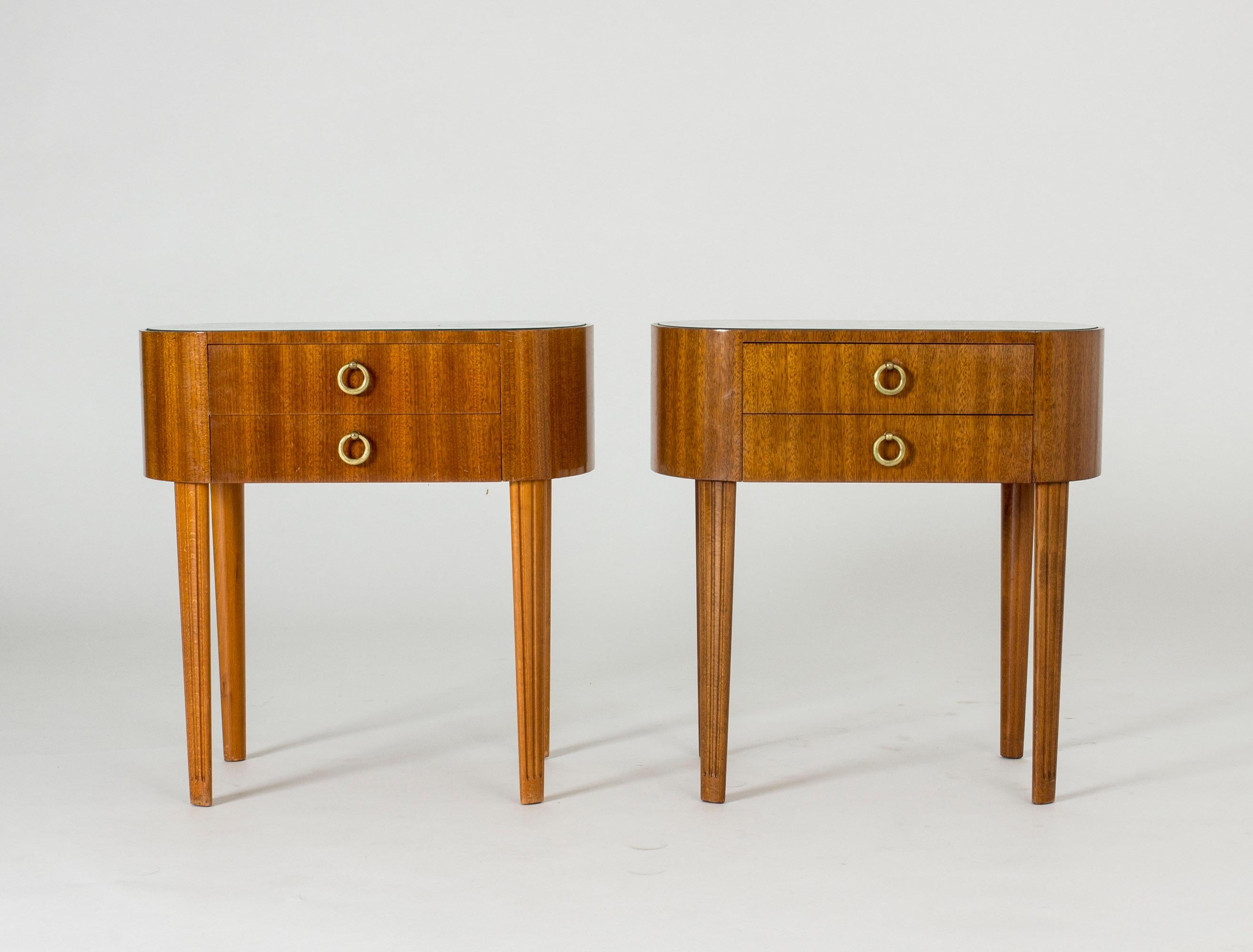 Pair of neat Swedish Grace bedside tables with round corners, made with striped mahogany veneer and tapering legs with embossed stripes. Glass table top and round brass handles.