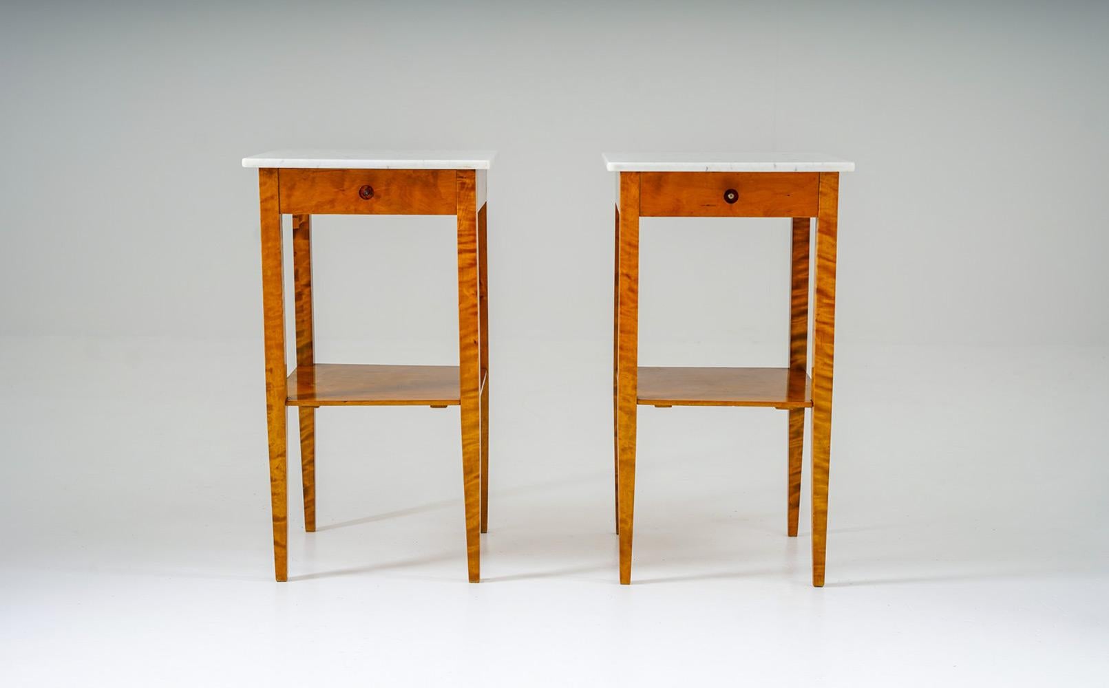 Paire de tables de nuit suédoises des années 1930, fabriquées en bouleau richement figuré, avec d'élégants pieds élancés et des plateaux en marbre blanc. Chaque support est équipé d'un petit tiroir et d'une étagère inférieure ouverte, combinant un