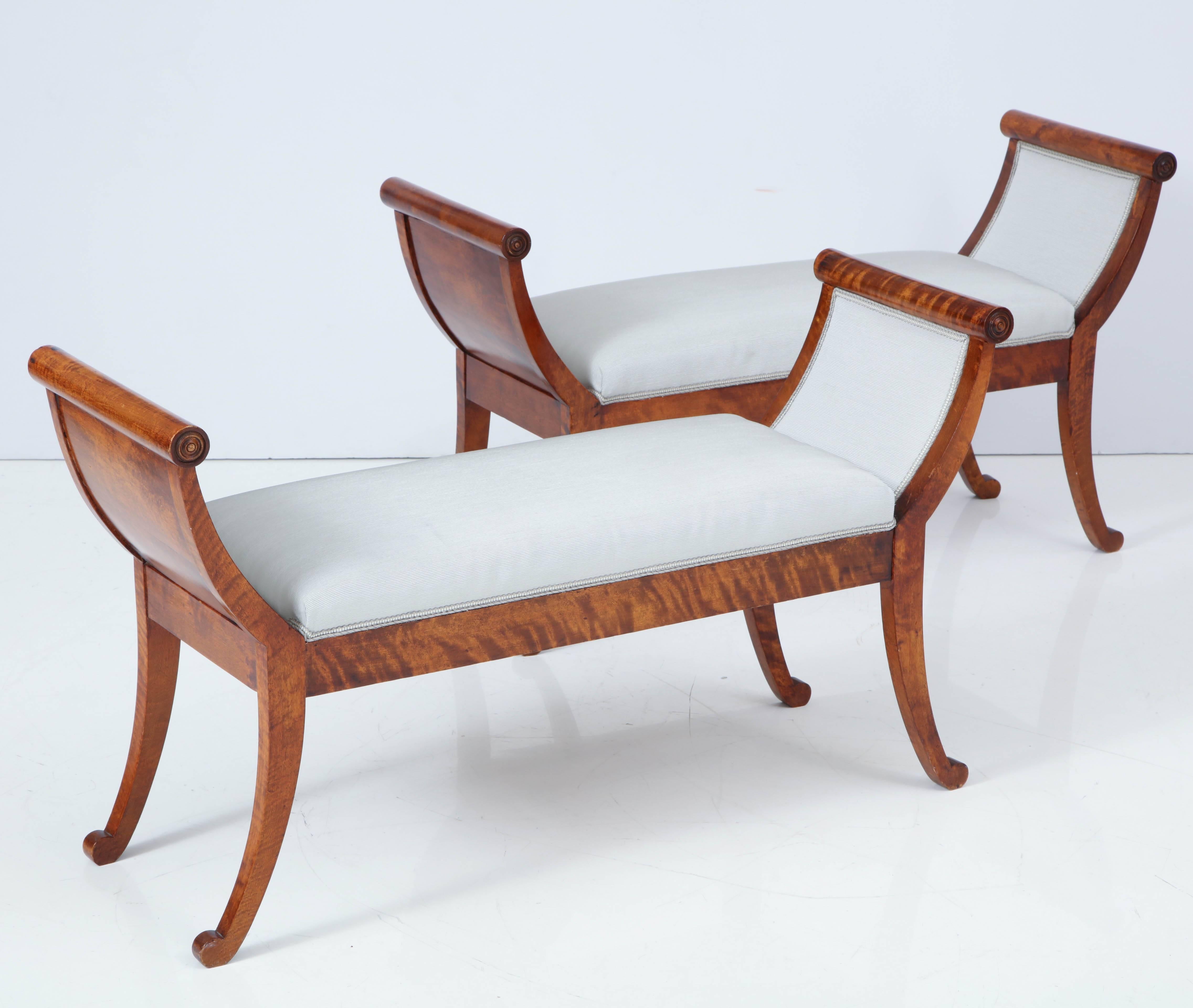 A pair of Swedish birchwood long or window benches, mid-19th century, the upholstered seats between outcurved armrests with carved roundels, raised on sabre legs ending with scrolls.
 