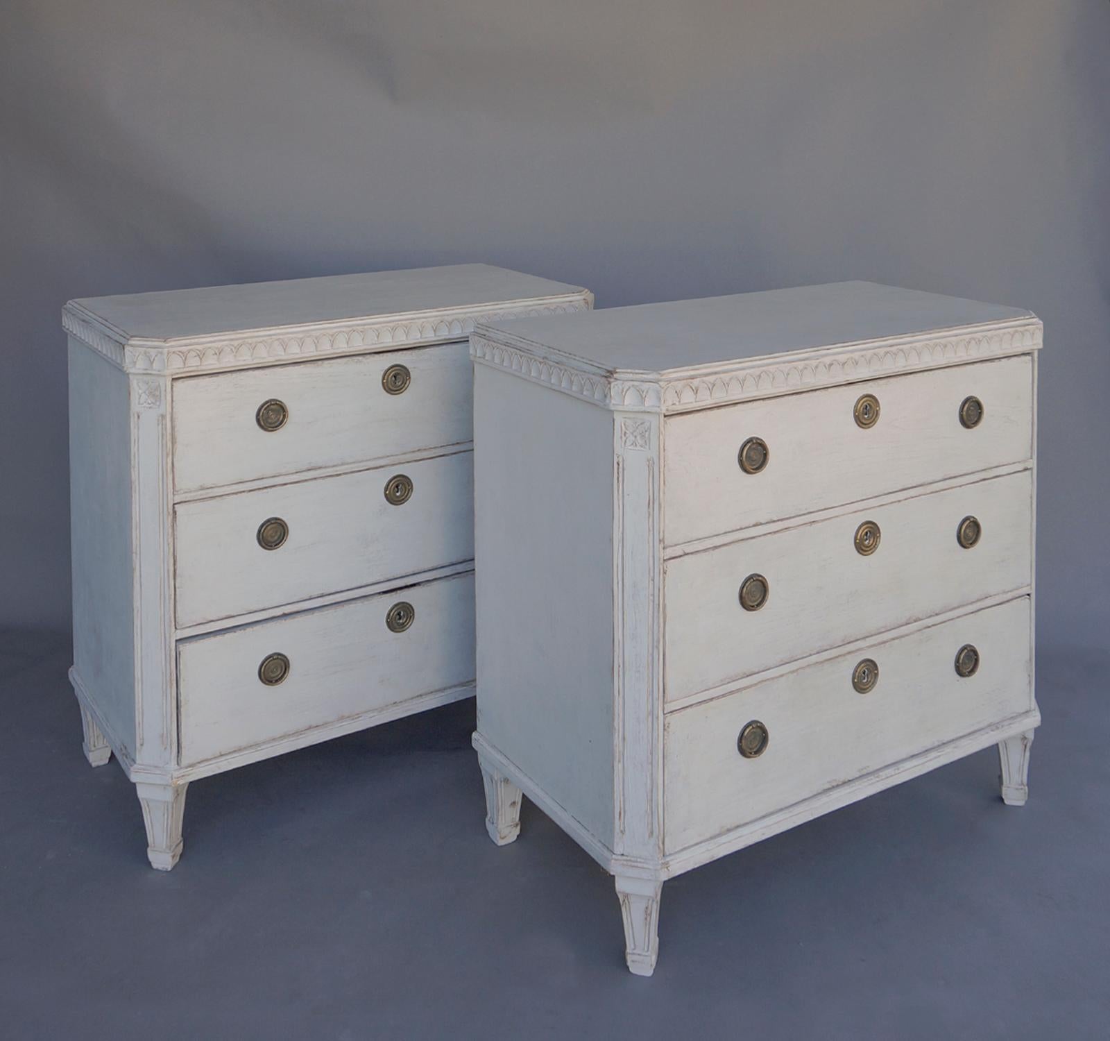 Pair of commodes, Sweden circa 1880, in the Gustavian Style. Shaped top with lamb’s tongue molding and canted corners with incised rosettes at the top. Brass hardware on the three drawers, later paint, and a repair to the back of one commode.