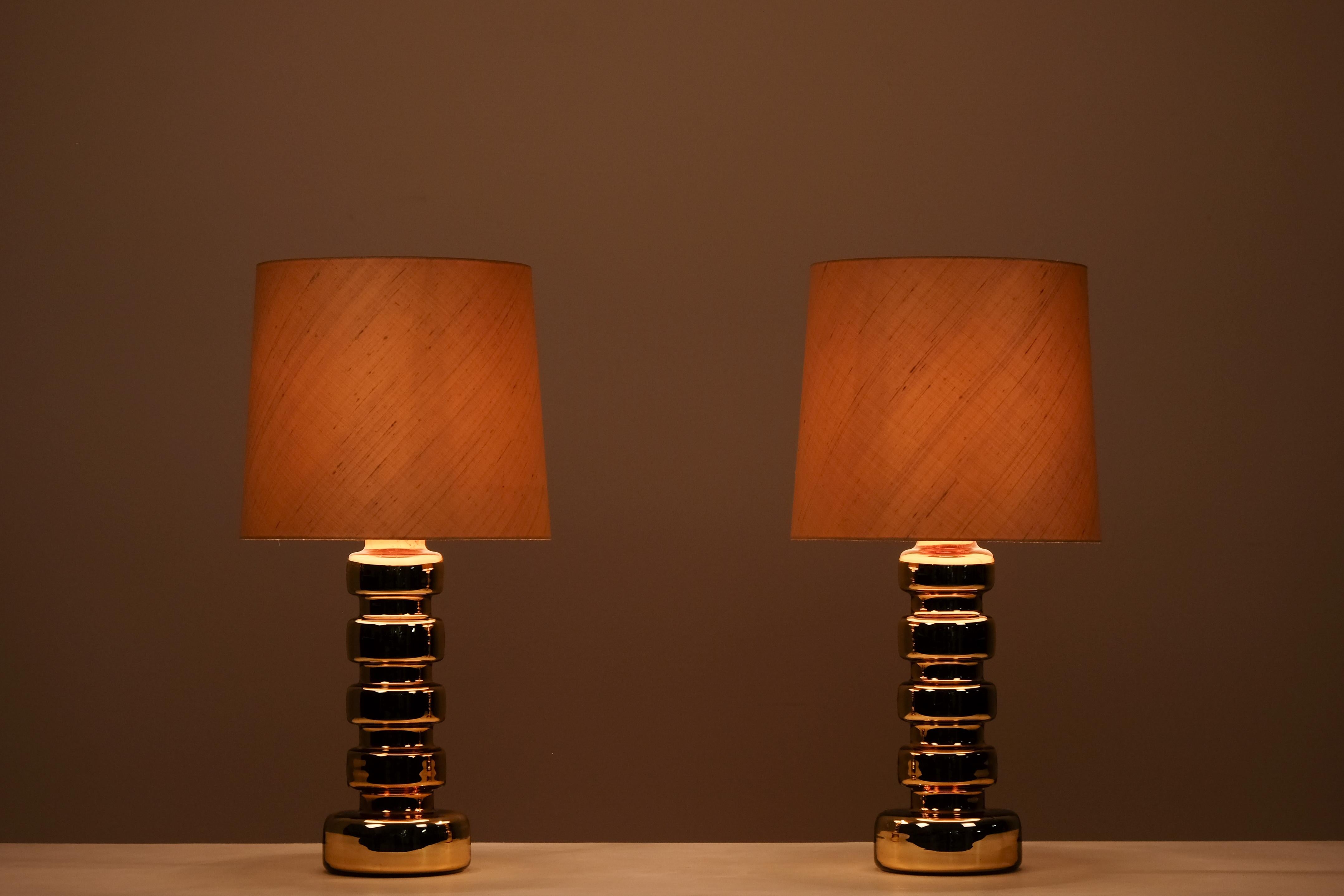Aporta calidez y carácter a tu espacio con este llamativo par de lámparas de mesa, fabricadas en Suecia en la década de 1960. Con unas esculturales bases de cristal dorado y un diseño brillante y escalonado, estas lámparas son auténticos reclamos