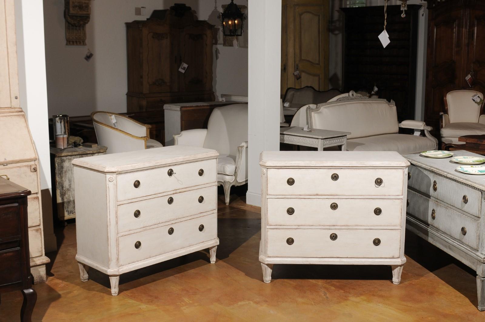 A pair of Swedish cream painted Gustavian style three-drawer commodes from the late 19th century, with canted side posts and tapered feet. Born in Sweden during the last quarter of the 19th century, each of these wooden chests features the stylistic
