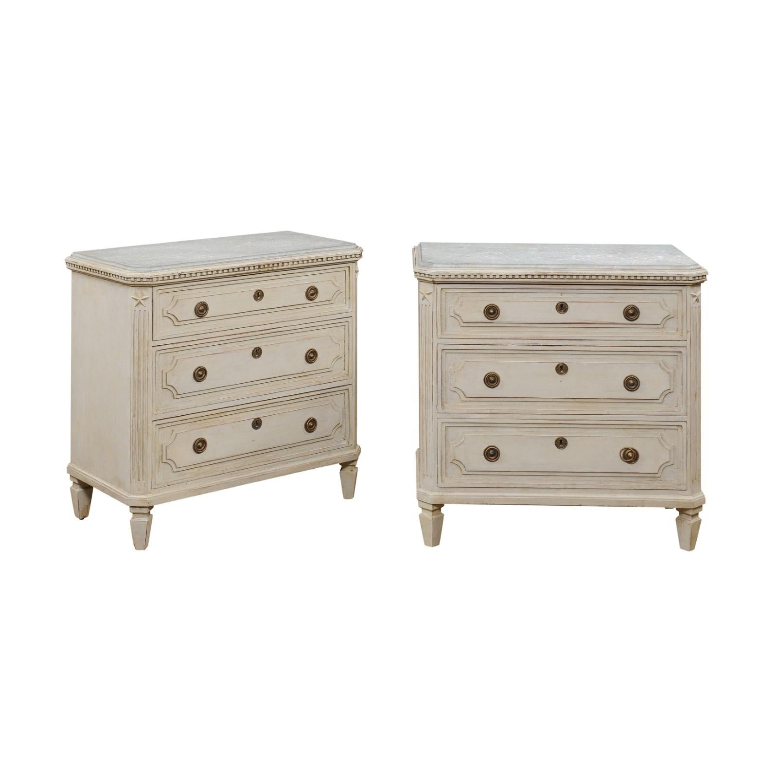 A pair of Swedish Gustavian style painted wood chests from the late 19th century, with marbleized tops, three graduated drawers, dentil molding, carved stars and fluted side posts. Created in Sweden during the last quarter of the 19th century, each